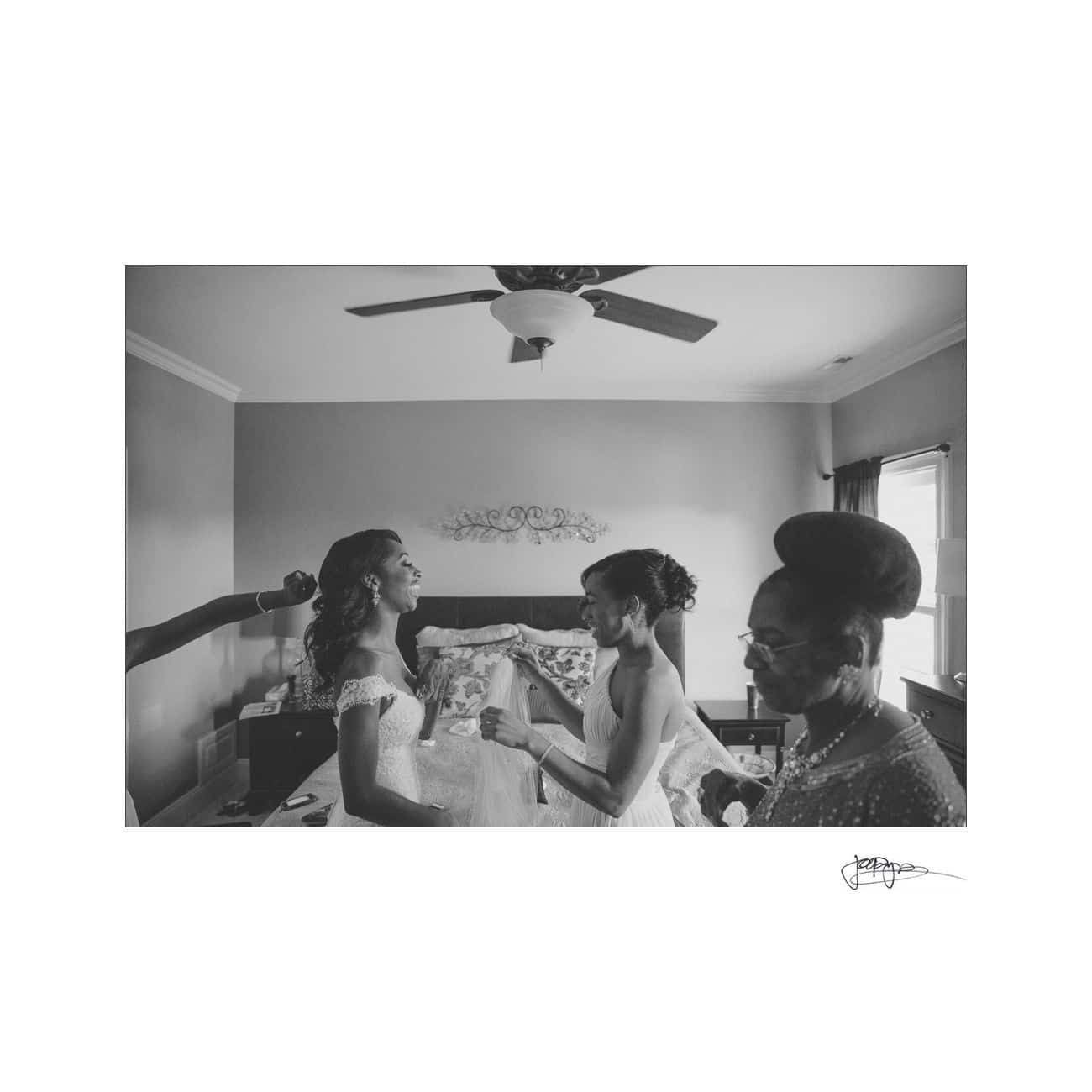 A black and white photo of three women in a bedroom. One woman, wearing a dress, stands as another adjusts a strap, while the third watches with a smile. A ceiling fan and curtained window are in the background.