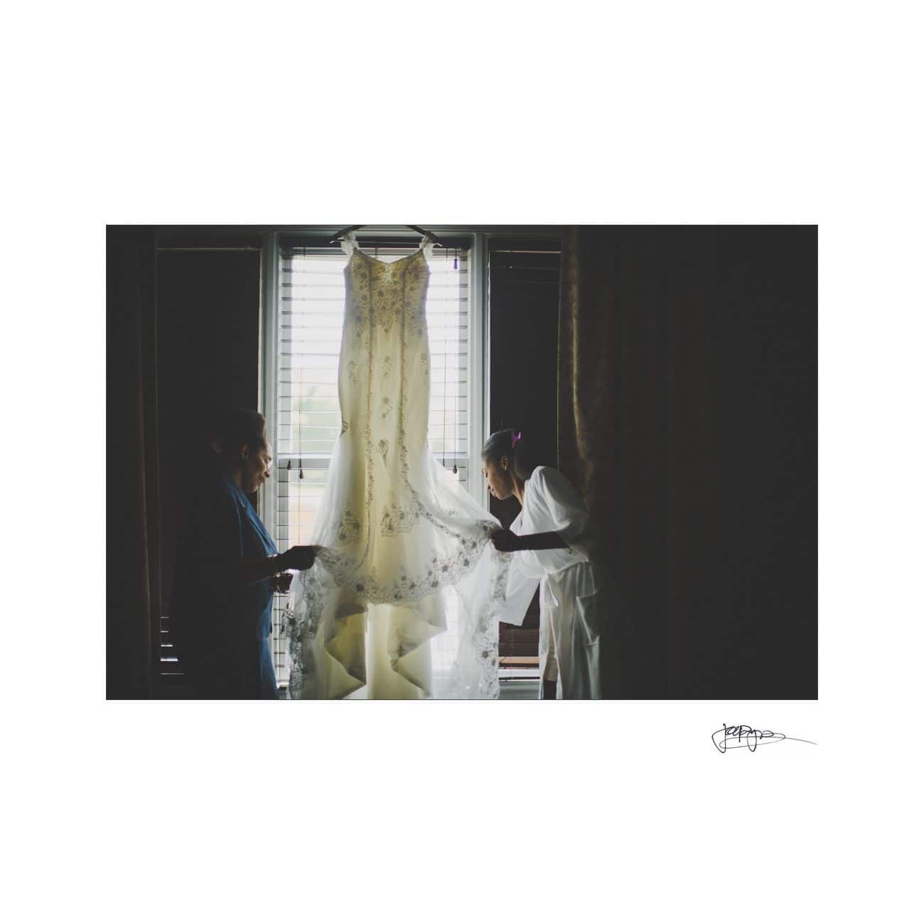 Two people holding up a wedding dress in front of a window, with light streaming through. The silhouette of the dress and the individuals are visible against the bright backdrop, creating an intimate and serene atmosphere.