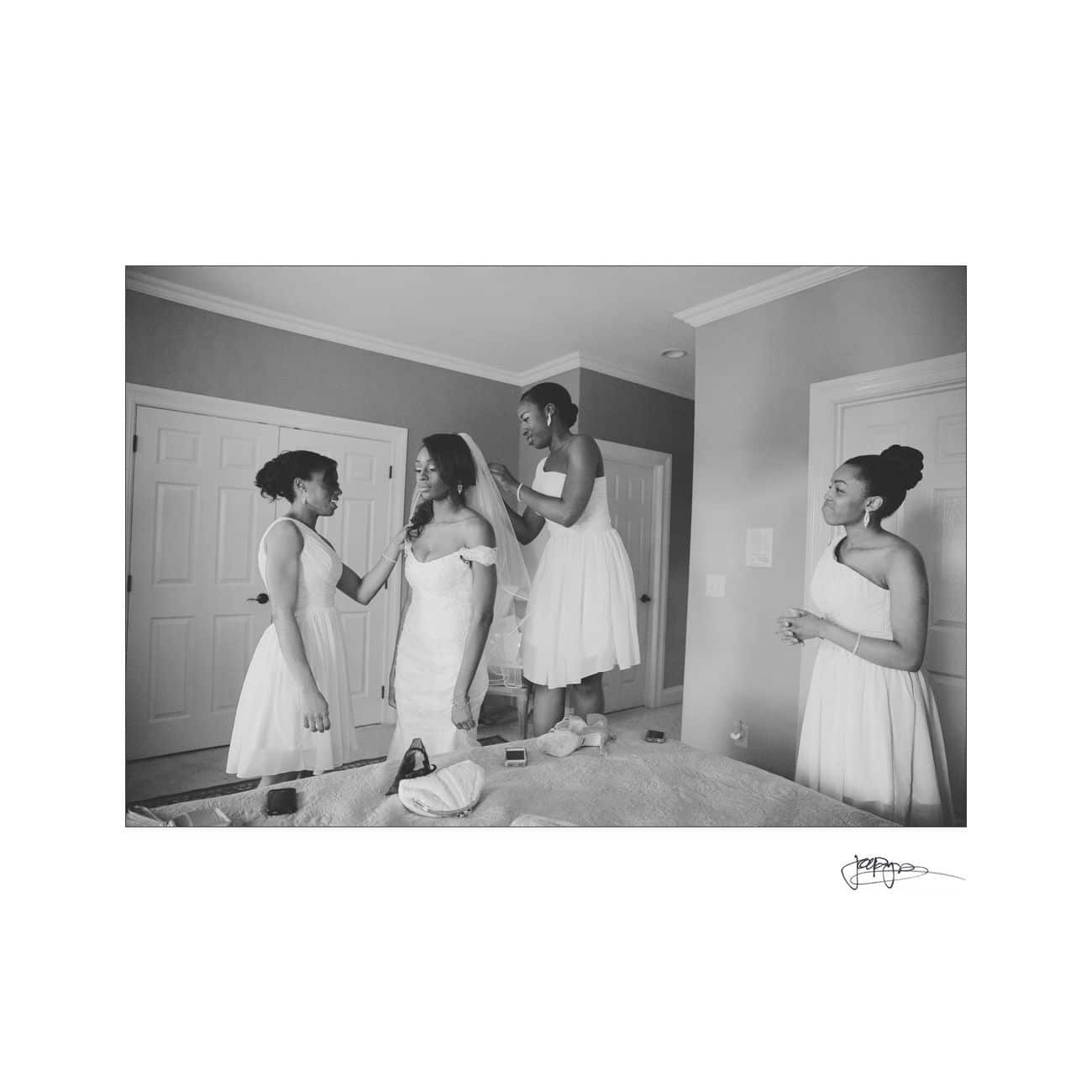 A bride in a strapless gown is surrounded by three bridesmaids in sleeveless dresses. One bridesmaid stands on a bed adjusting the brides veil, while another holds her hand. They are in a room with closed double doors. The scene is black and white.