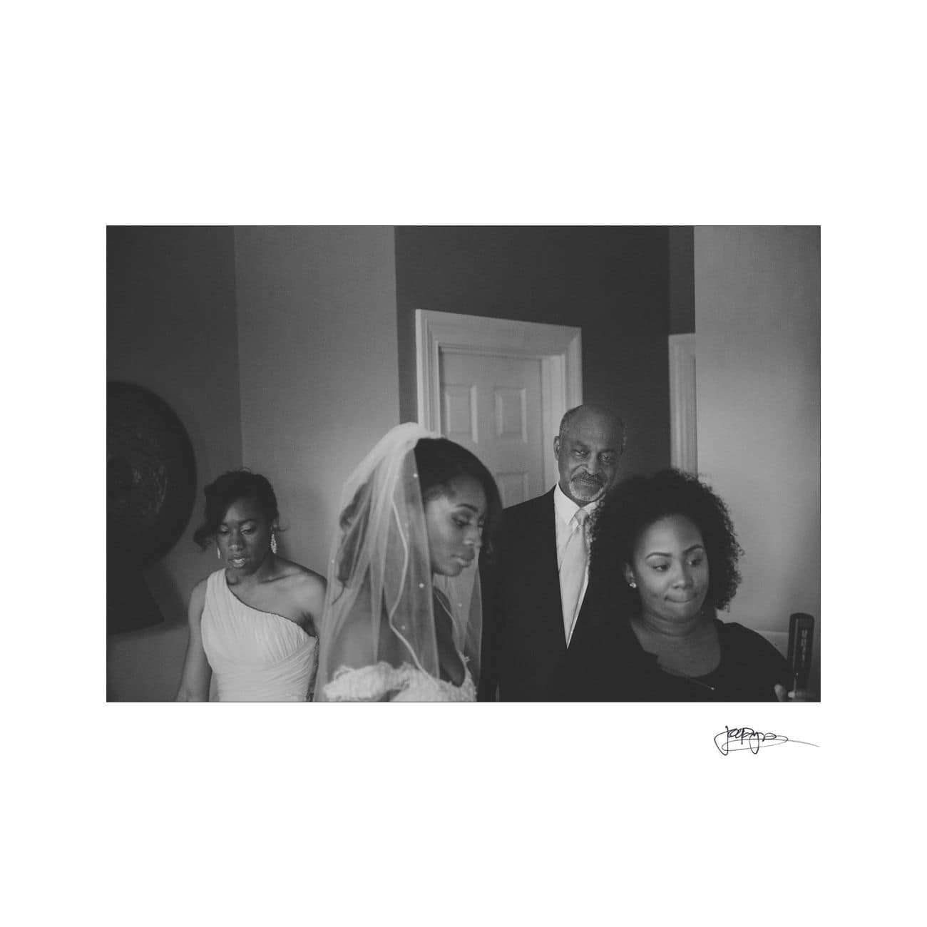 A black and white photo of a bride wearing a veil surrounded by three people. One is an older man and two are women dressed in formal attire. They appear to be in a room, perhaps gathering before a wedding ceremony.