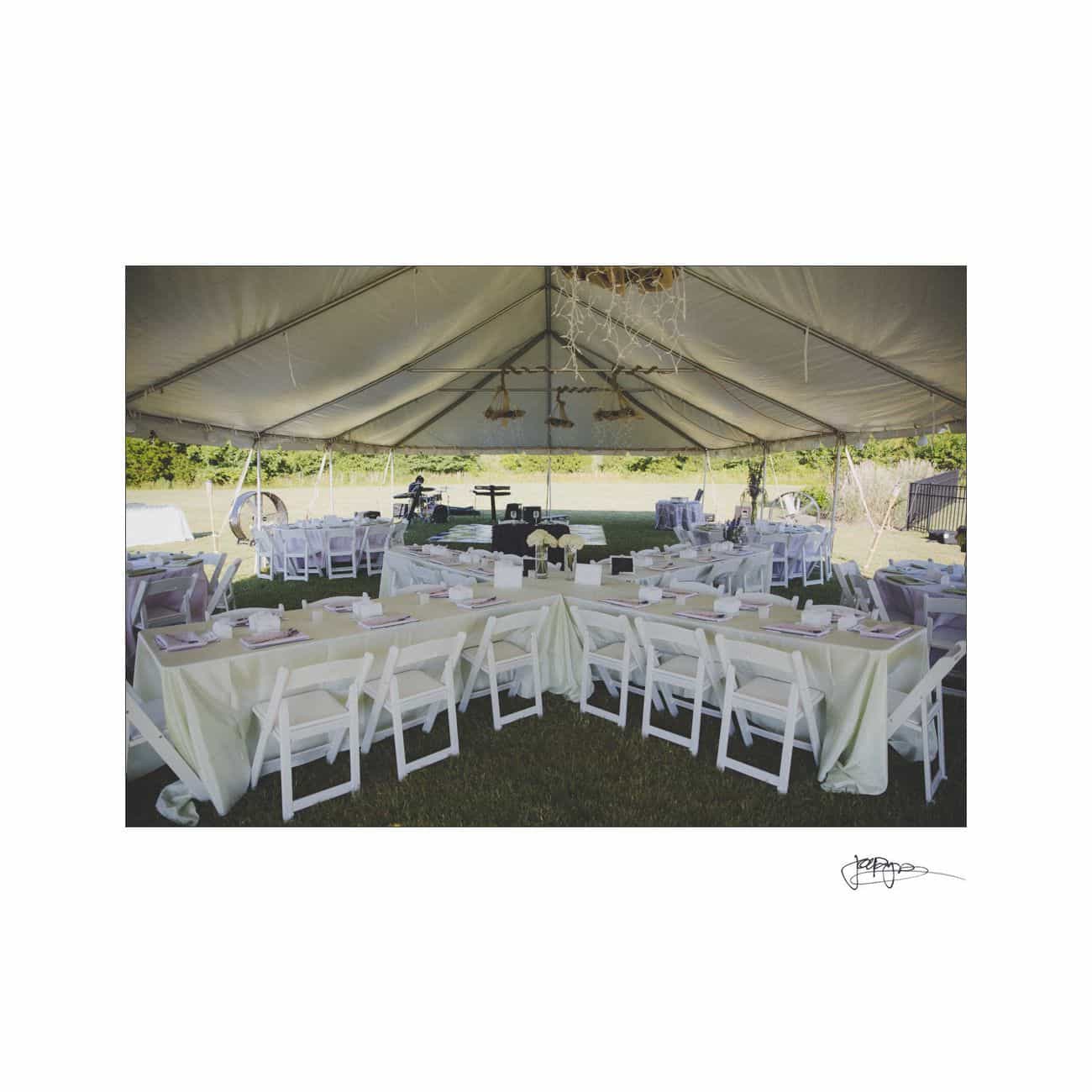 A large white tent set up outdoors with multiple tables and chairs arranged for an event. The tables are covered with white tablecloths and have place settings. Decorations hang from the ceiling. The scene is surrounded by greenery.