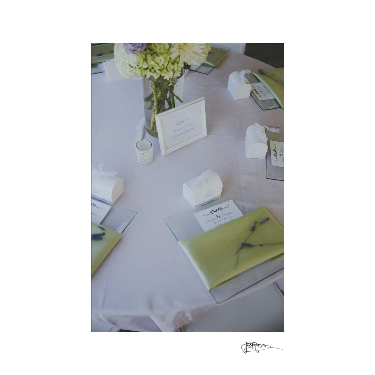 Elegant table setting with a white tablecloth, green napkins, and small lavender sprigs. Each place has a white gift box. A centerpiece with white and purple flowers sits in the middle, next to a framed sign.