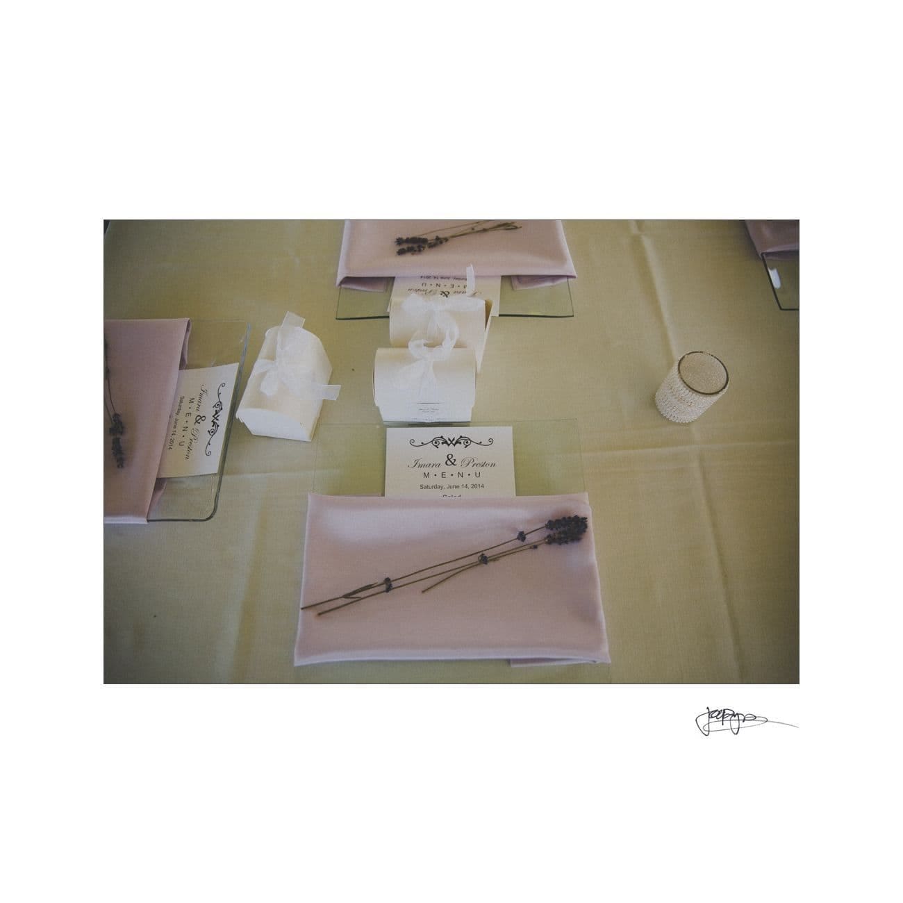 A table setting featuring a pink napkin on a glass plate, topped with sprigs of lavender. To the side, a small white box tied with a ribbon, a card with the names Kristine & Patrick, and a glass votive candle sit on a light-colored tablecloth.