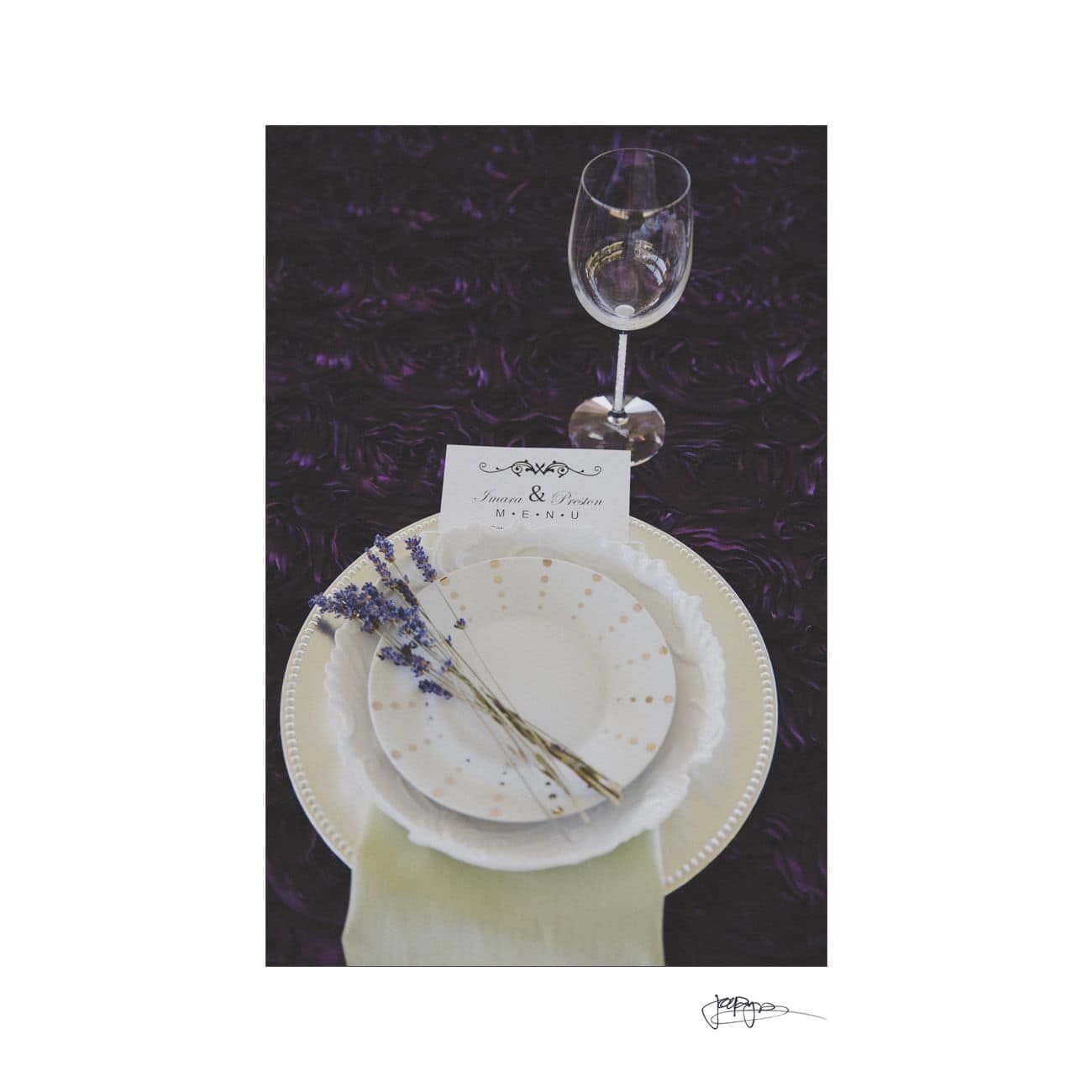 A table setting featuring stacked plates with a sprig of lavender on top, a menu card, and a green napkin. A wine glass stands nearby on a dark, textured tablecloth with a decorative pattern.