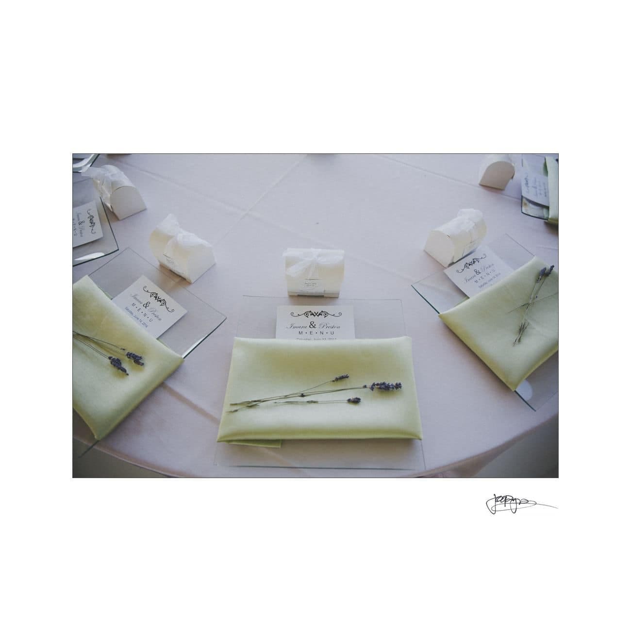 A table setting with pale yellow napkins on clear plates, adorned with sprigs of lavender. Each setting includes a small white box and a place card on a round white tablecloth.