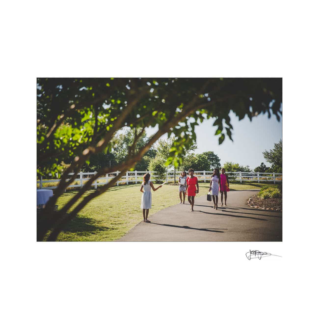 Four people are walking on a paved path surrounded by grass and trees. The image is framed by tree branches, and a white fence lines the grassy area. The figures are wearing colorful clothing on a sunny day.