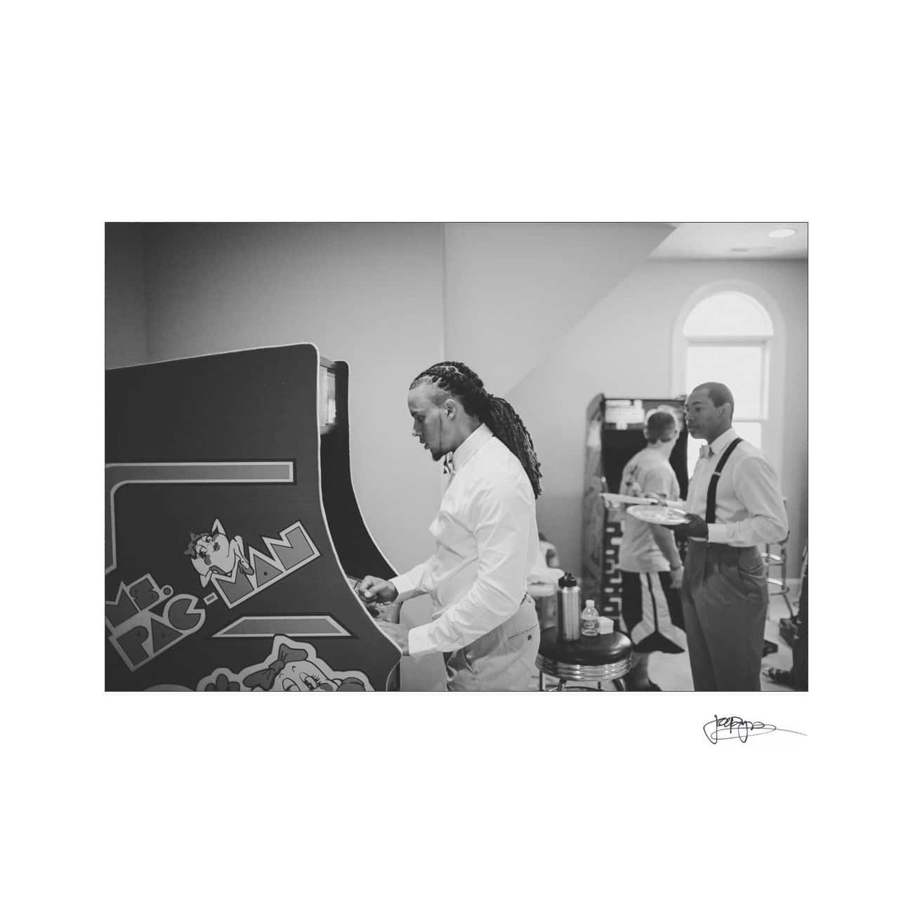 A black and white photo of a man playing a vintage arcade game. Another man nearby is holding a drink and watching. The room has a large window, and the lighting creates a soft atmosphere.