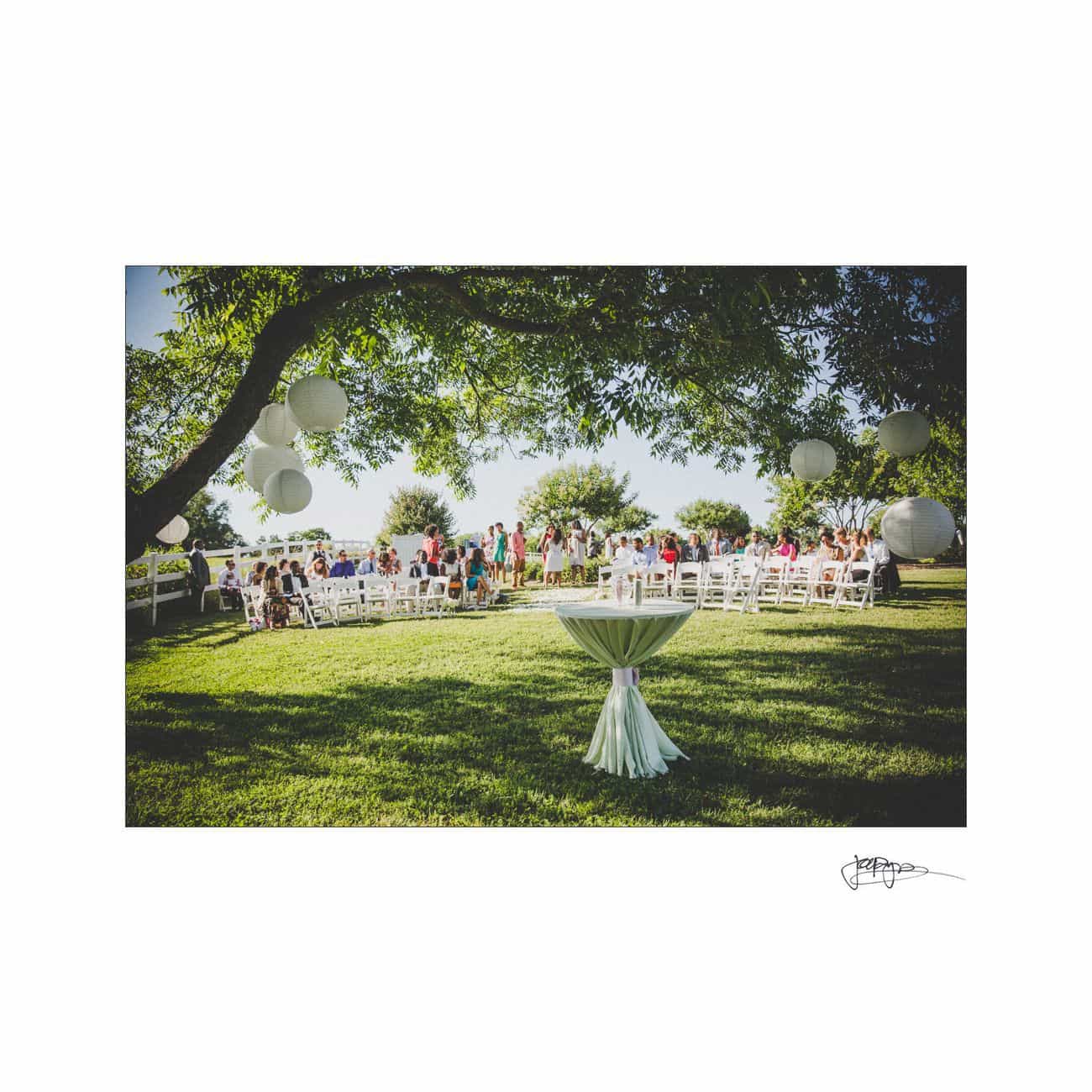 A wedding ceremony set outdoors under a tree adorned with paper lanterns. Rows of white chairs are arranged for guests, facing a white altar cloth draped over a small table. The scene is bright and sunny with a clear blue sky.