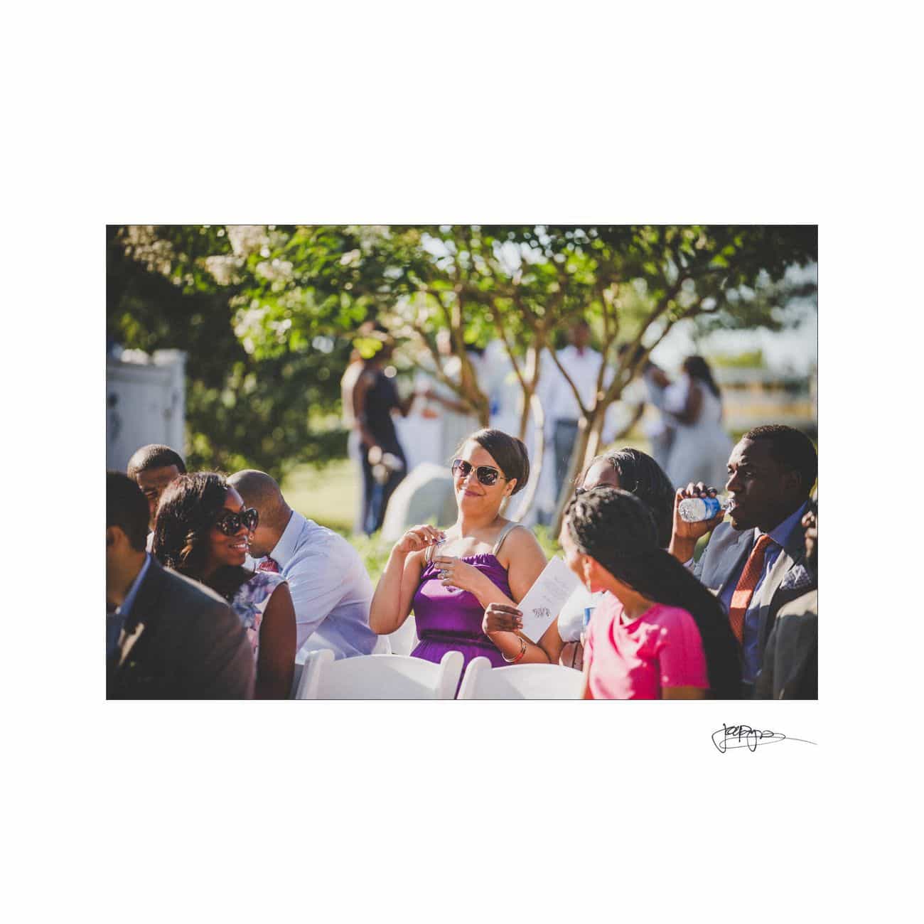 A group of people seated outdoors at an event. A woman in a purple dress and sunglasses smiles while holding a cup. Sunlight filters through the trees, creating a warm and cheerful atmosphere.