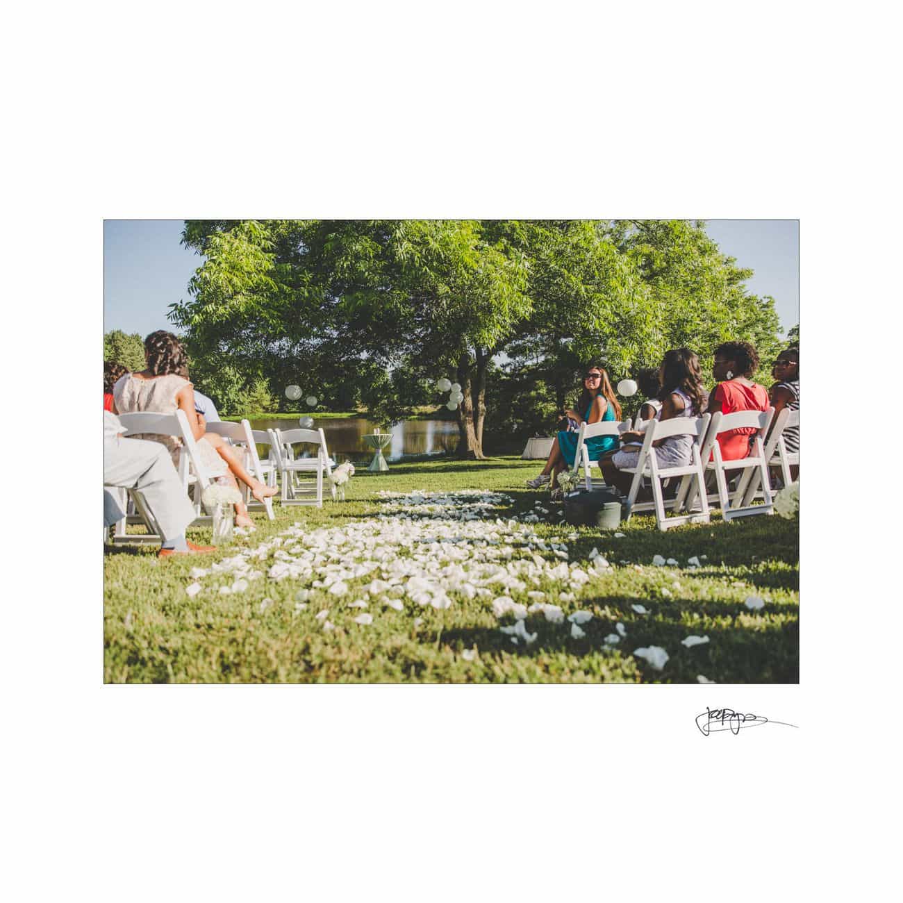 A serene outdoor wedding ceremony setup with white chairs arranged on either side of an aisle adorned with white petals. Guests are seated, and a large tree and a pond are visible in the background under a clear sky.
