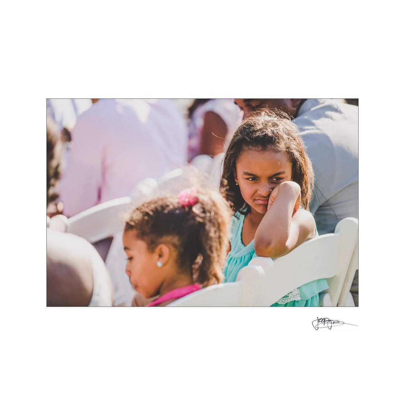 Two children seated outdoors at an event. The girl in the foreground is wearing a teal dress and is resting her head on her hand, looking pensive. Another child in a pink dress sits beside her, with her hair tied up. People are seated around them.