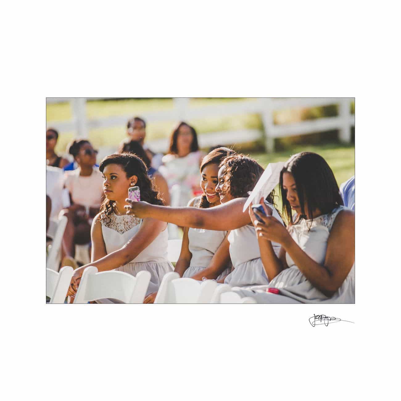 A group of bridesmaids in gray dresses sit on white chairs outdoors. One woman is holding a phone, possibly taking a photo. The background is a sunny day with guests seated on a grassy area.