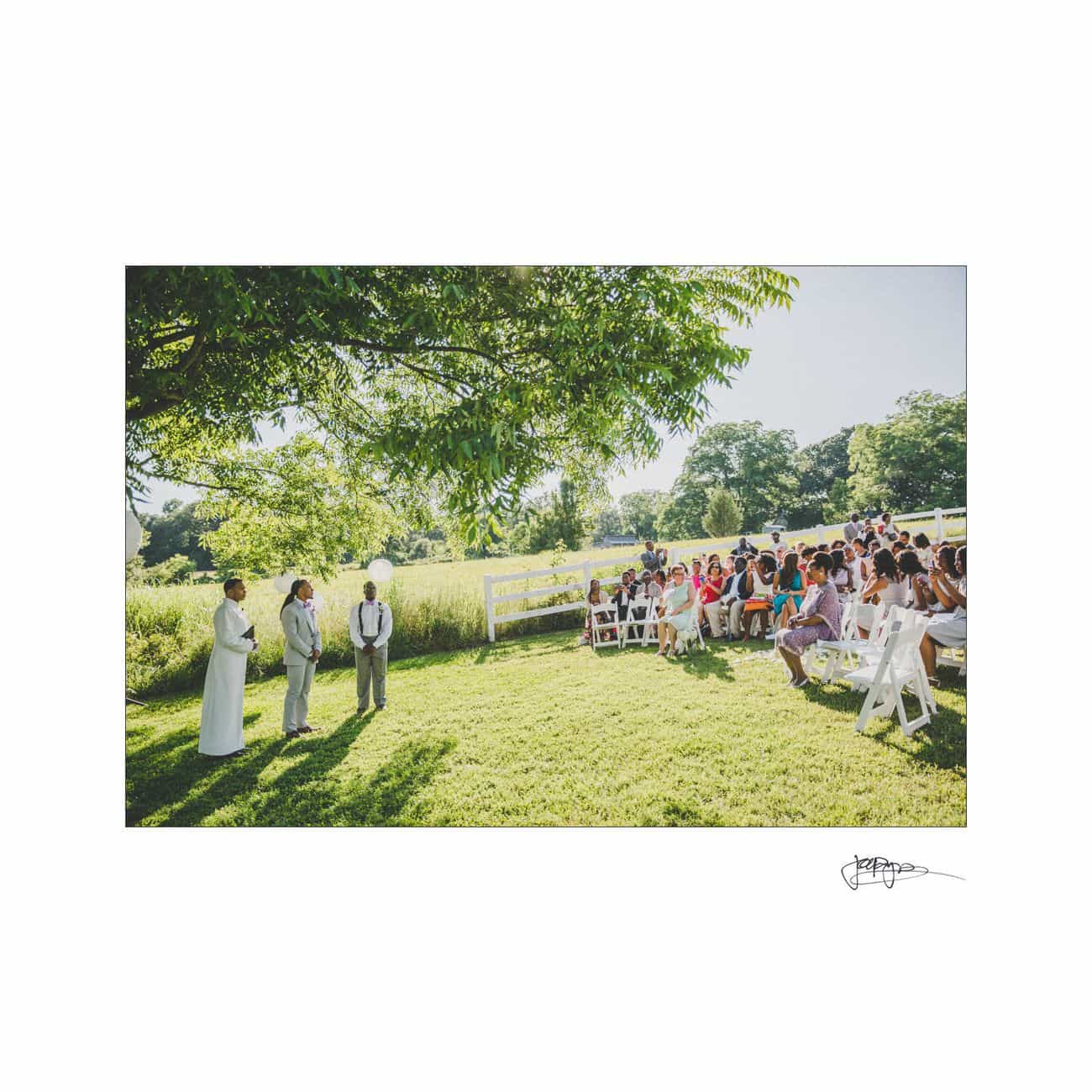 A sunny outdoor wedding ceremony with guests seated on white chairs. A couple stands with an officiant under a large tree in a grassy field, surrounded by white fences and lush greenery.