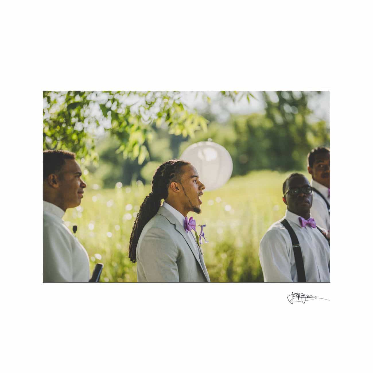 A groom in a light gray suit with a purple bow tie stands among groomsmen outdoors. The scene is decorated with a large white balloon against a backdrop of soft greenery under natural sunlight.