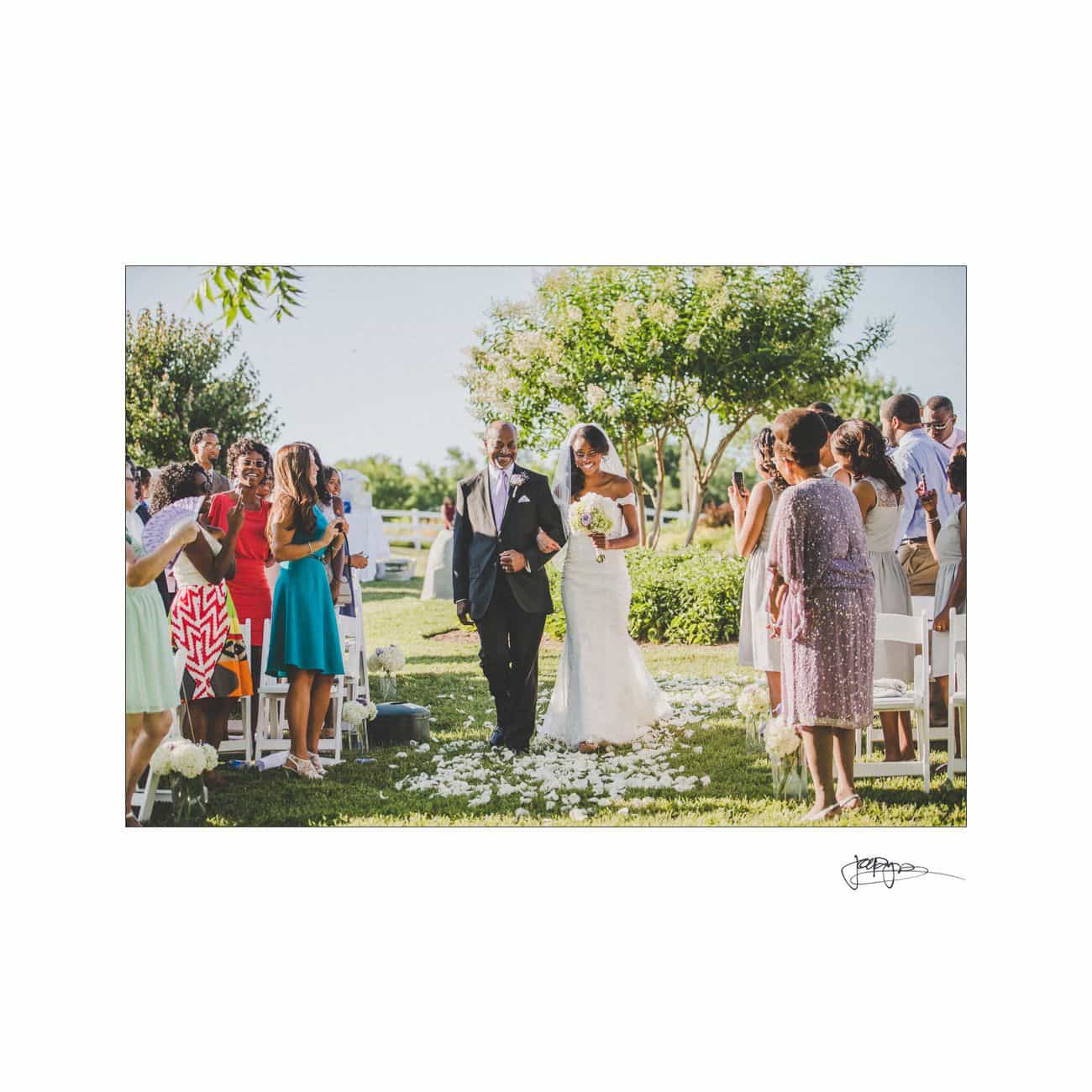 A bride in a white dress and veil walks down an outdoor aisle with an older man. Guests on either side stand among white chairs, clapping and celebrating. The aisle is adorned with white rose petals, and trees and grass surround the scene.