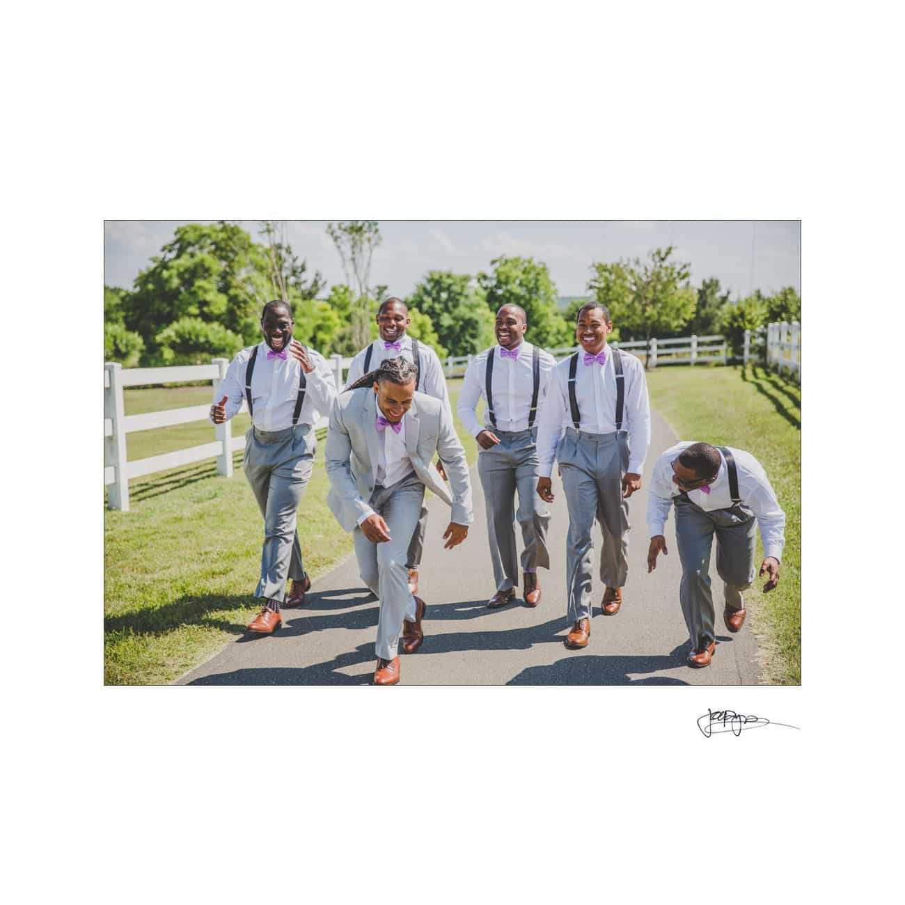 A group of five groomsmen in light gray suits and suspenders are laughing and playing around while walking on a paved path beside a wooden fence and green fields. The sky is clear and sunny.