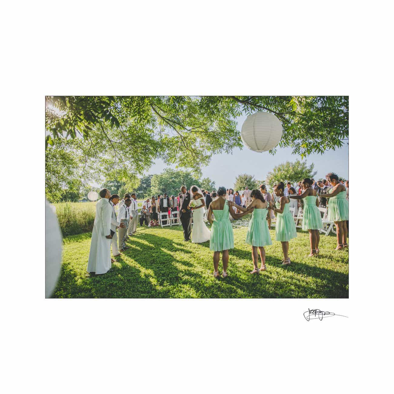 A wedding ceremony outdoors with a group of bridesmaids in mint dresses and groomsmen in light suits forming a circle. Paper lanterns hang from tree branches above. Guests are seated on white chairs under sunny skies.