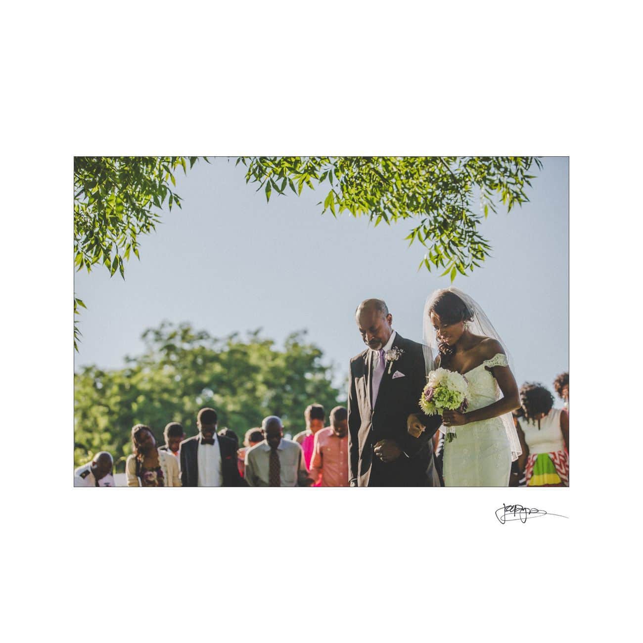 A bride in a white dress and a man in a suit walk together outdoors during a wedding ceremony. They are surrounded by guests, with green trees and a clear sky in the background.