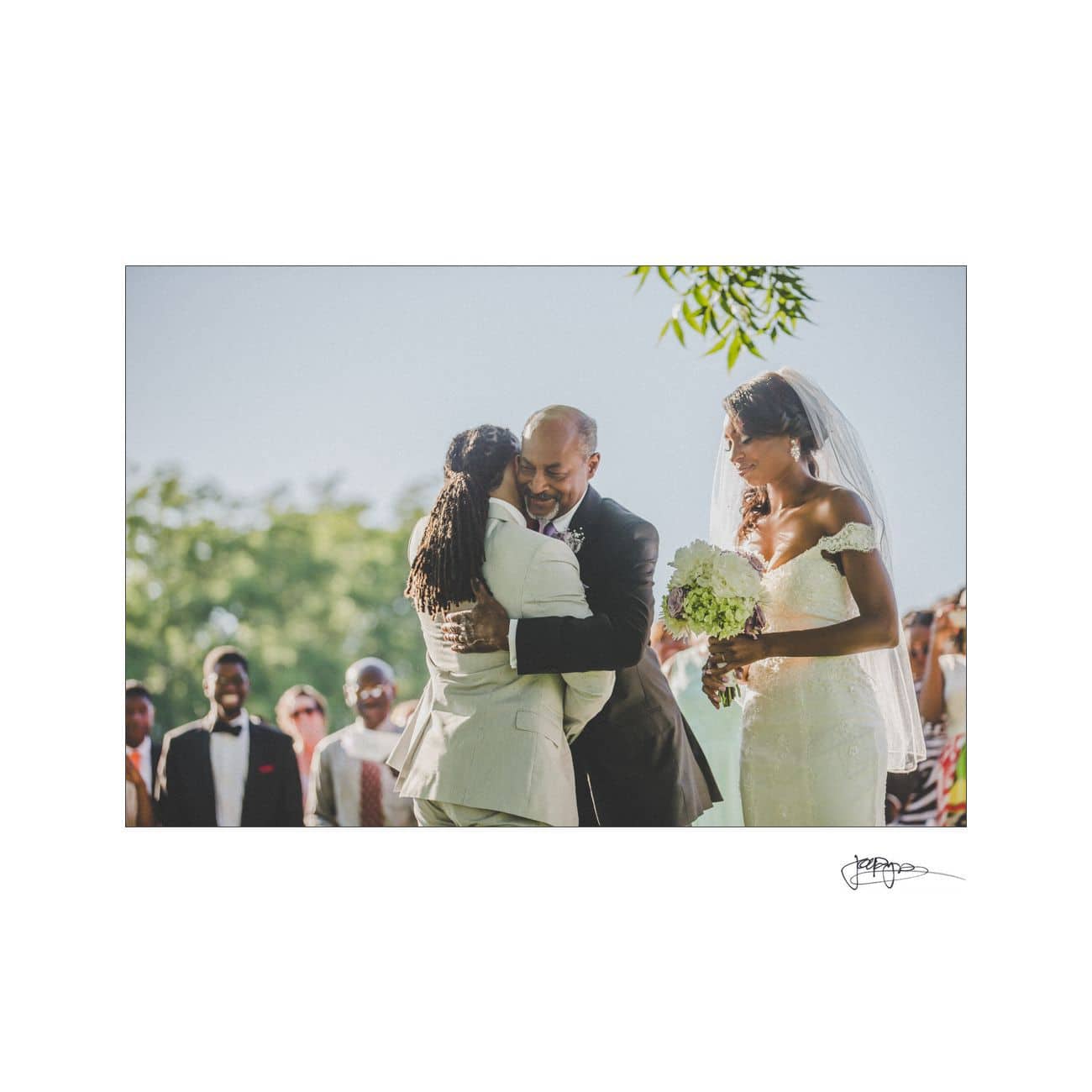 A bride in a white dress stands smiling at a couple hugging. The groom, in a suit, embraces a person wearing a white suit. They are outdoors, surrounded by smiling guests. The atmosphere is joyful and celebratory.