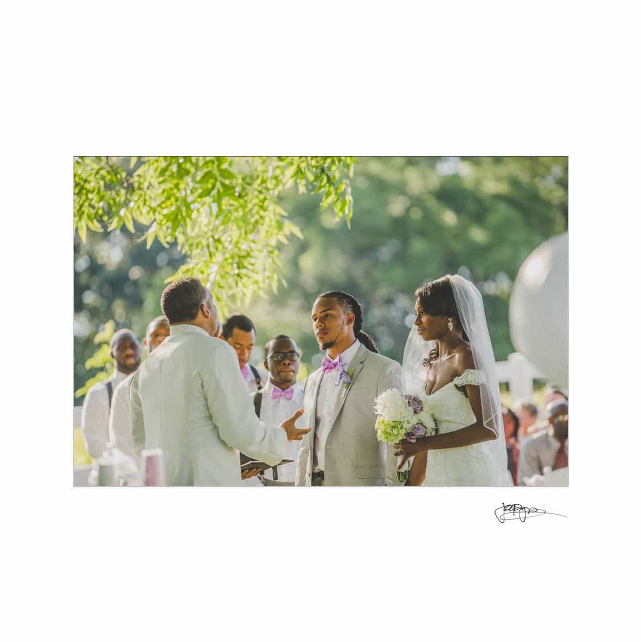 A bride and groom stand together during an outdoor wedding ceremony. The groom wears a light suit and purple bow tie, while the bride holds a bouquet and wears a veil. The officiant faces them, with guests watching in the background.
