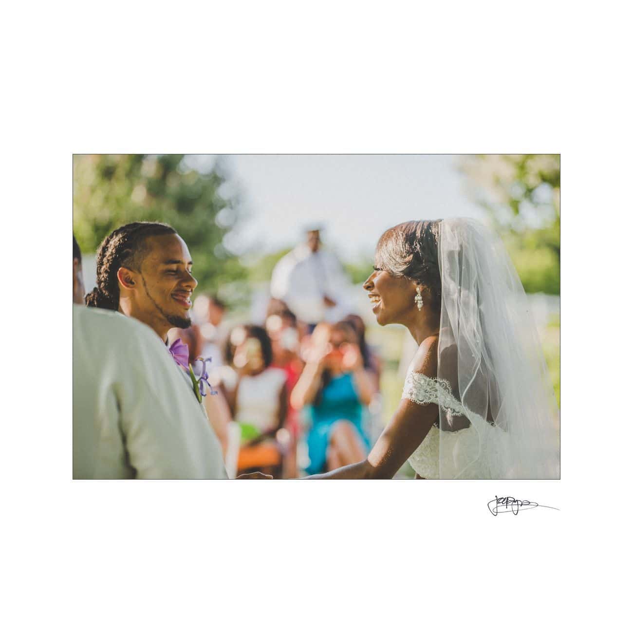 A bride and groom sit facing each other, smiling during an outdoor wedding ceremony. The bride is wearing a veil and holding a bouquet, while the groom is in a suit. Guests are seated in the background with greenery surrounding the area.