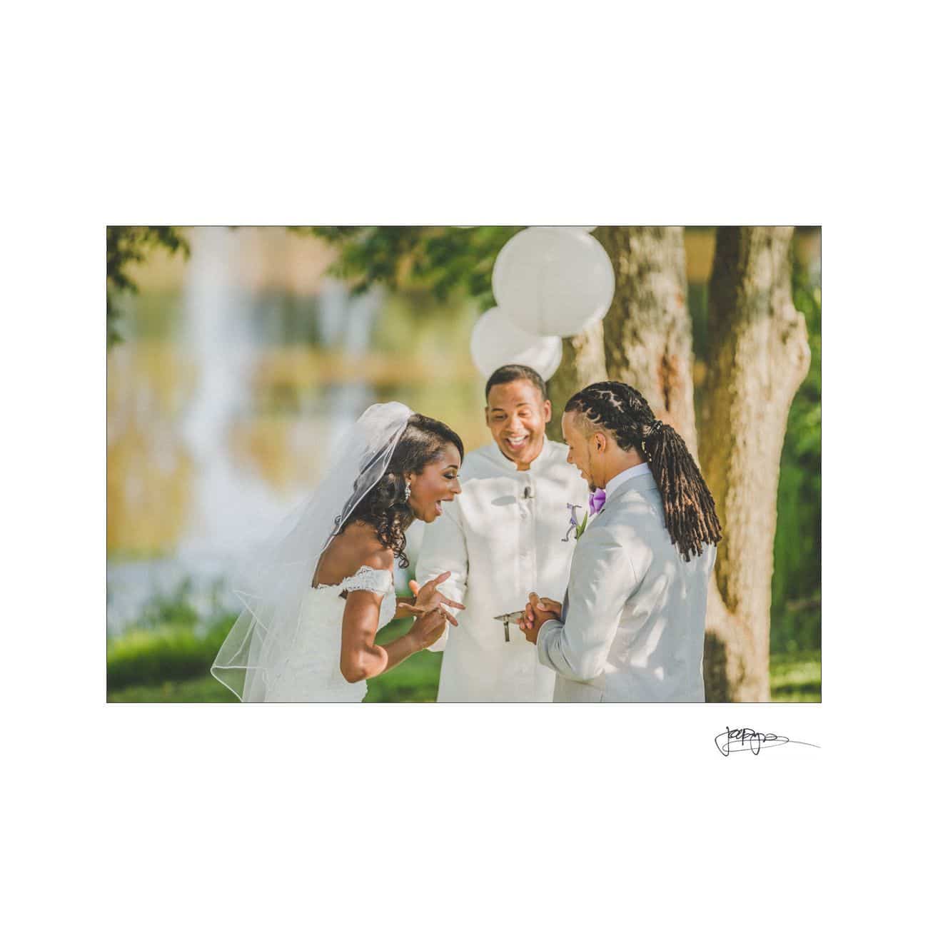 A bride and groom exchange rings during an outdoor wedding ceremony. They are smiling and laughing, standing under trees with white balloons in the background. The officiant, also smiling, stands behind them.