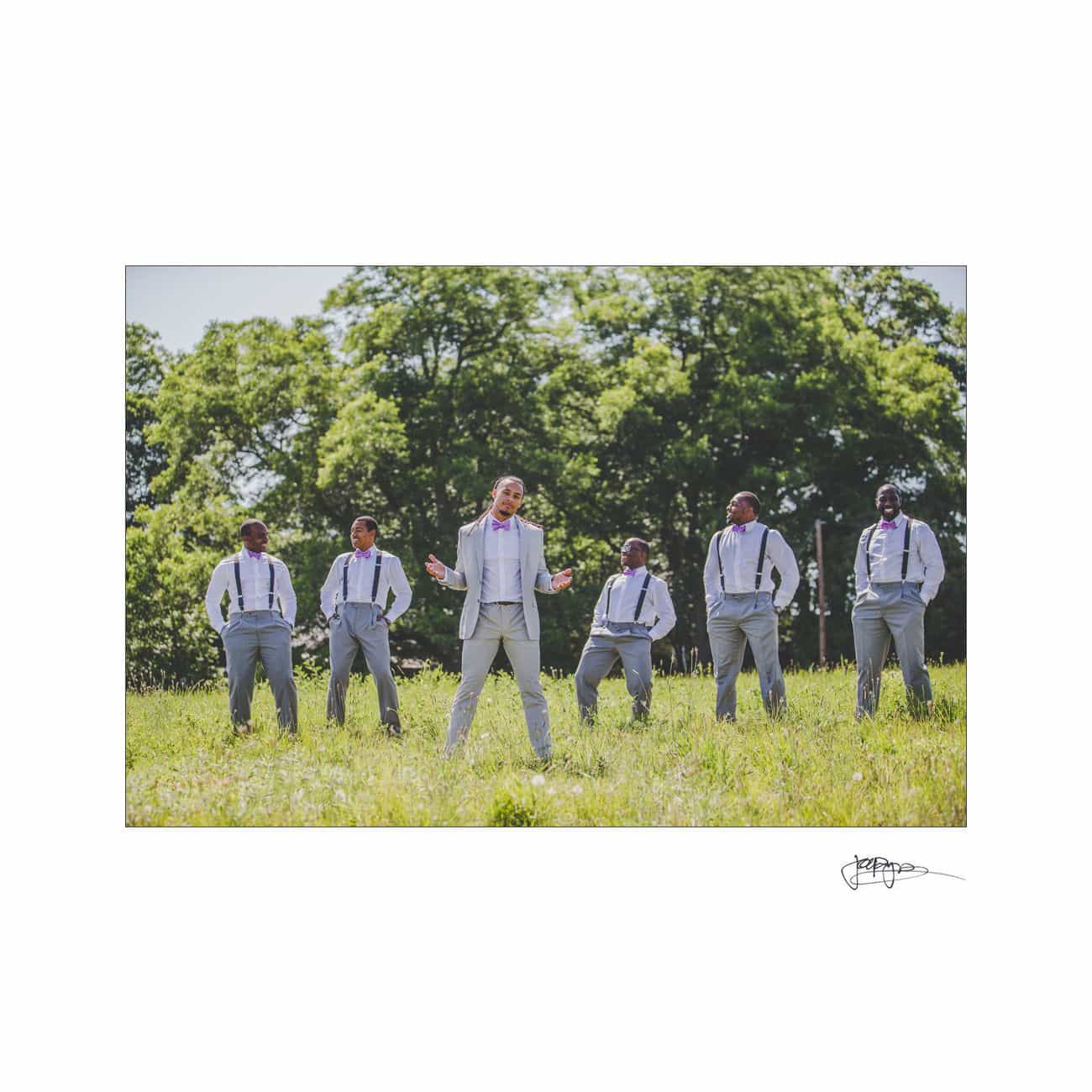 A group of six men in gray suits with suspenders stand in a grassy field. Five men stand in the background, while one man is in the foreground. Trees and a clear sky are visible in the background.