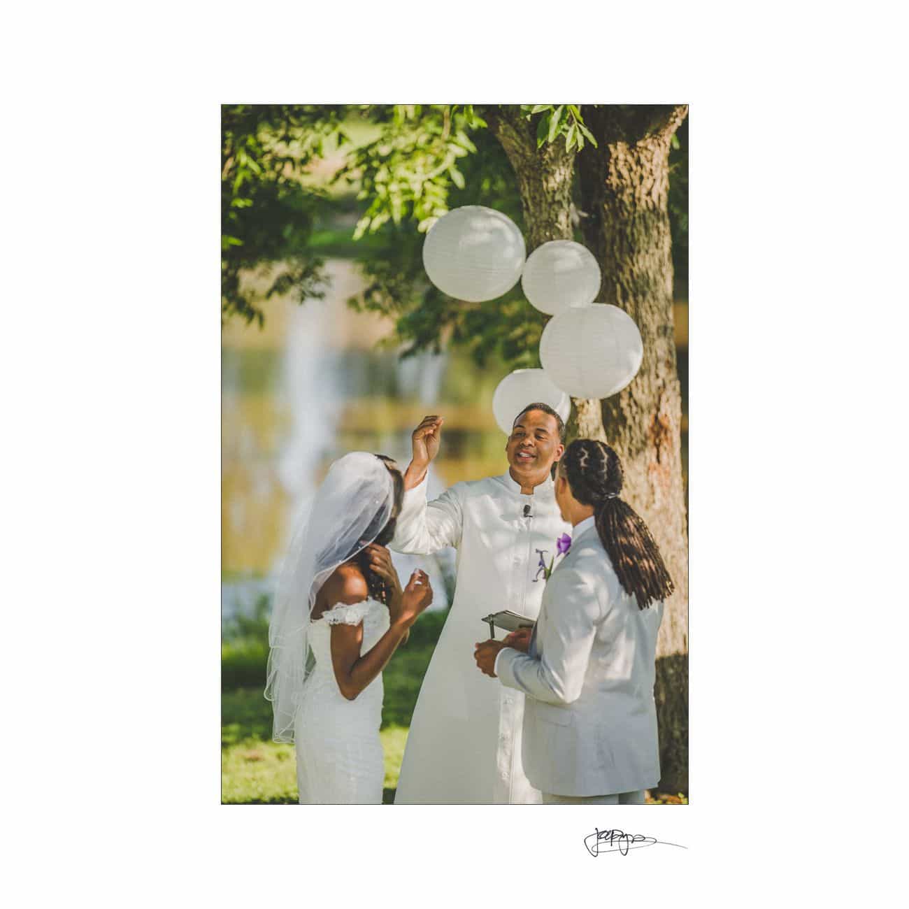 A couple is getting married outdoors under a tree with white lanterns hanging nearby. The bride is wearing a white dress and veil, the groom a white suit, and an officiant stands with them. A lake is visible in the background.