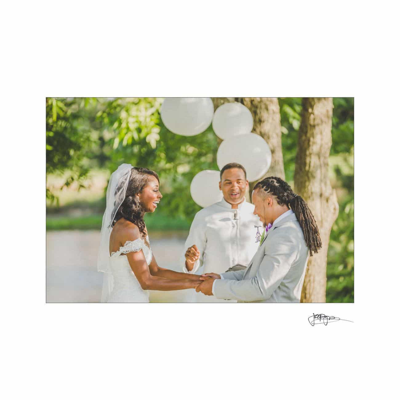 A joyful couple stands hand in hand during their outdoor wedding ceremony, surrounded by nature. The bride wears a white gown and veil, and the groom wears a light suit. A person officiates the ceremony, smiling, with white balloons in the background.