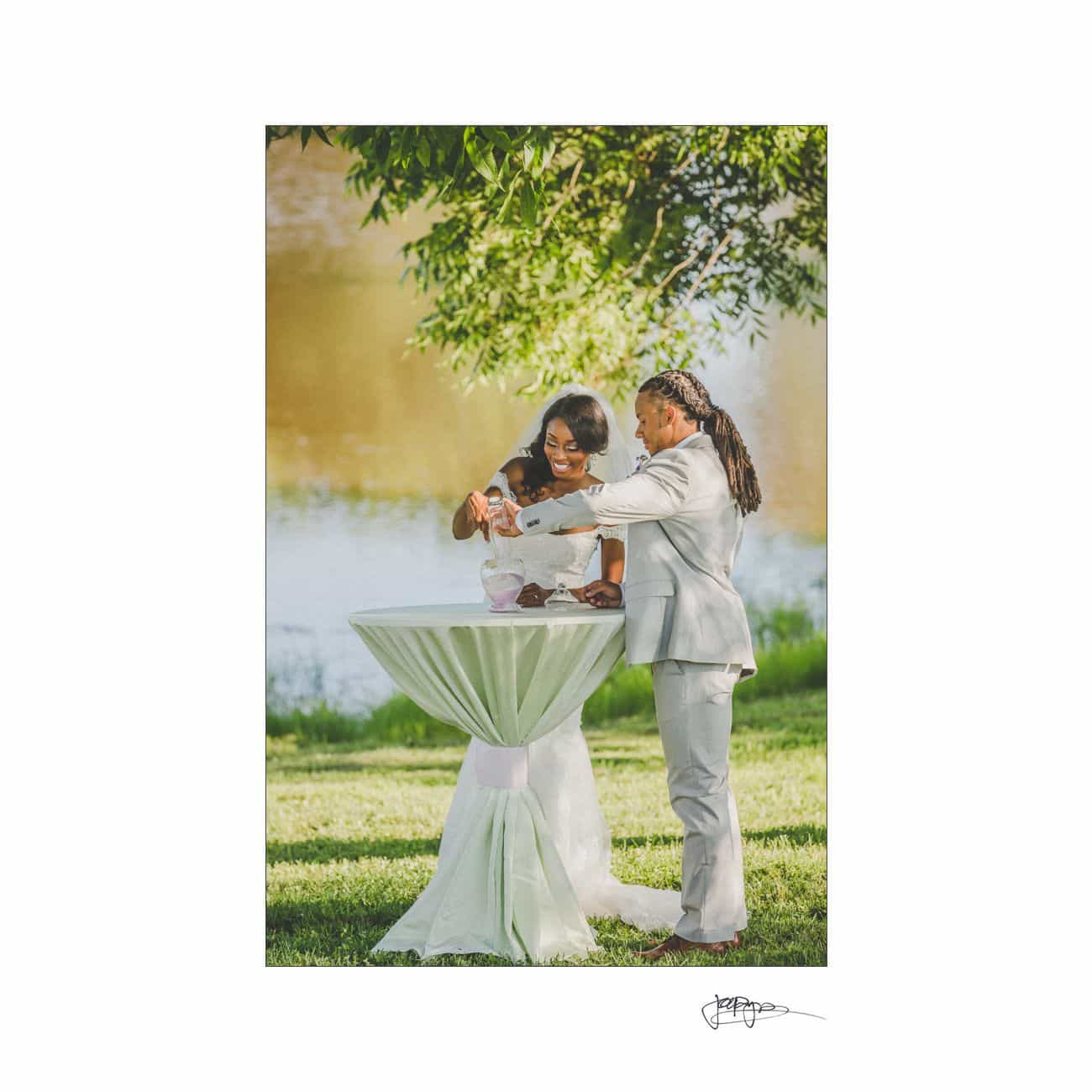 A couple in formal attire stands by a tall, round table outdoors near a body of water. They are helping a baby hold a white sippy cup. Lush greenery surrounds them, and the scene conveys a joyful, family-oriented moment.