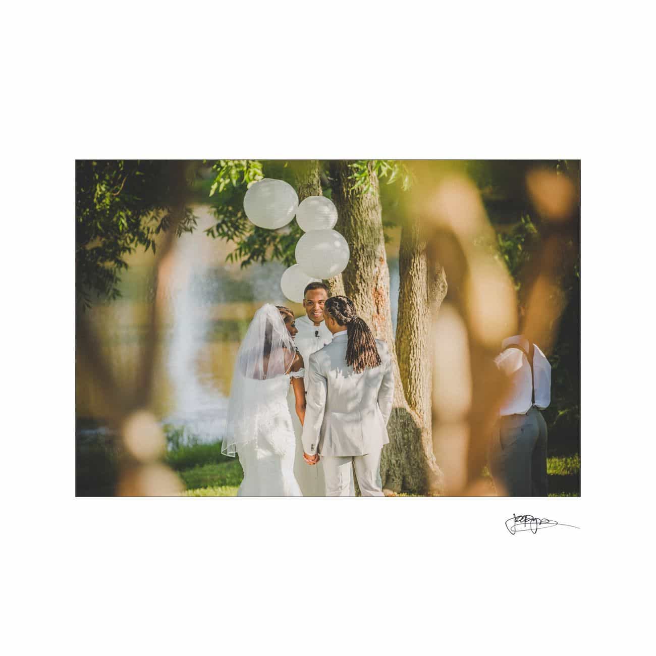 A bride and groom hold hands during an outdoor wedding ceremony, facing the officiant. They are surrounded by greenery and several white balloons, creating a serene and intimate atmosphere.