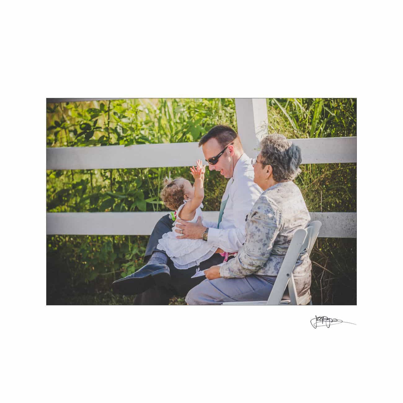A man sat on a white chair holding a toddler in a white dress, while an older woman in a floral shirt sits beside them. They are outdoors, near a white fence, with greenery in the background. The toddler is reaching up animatedly.