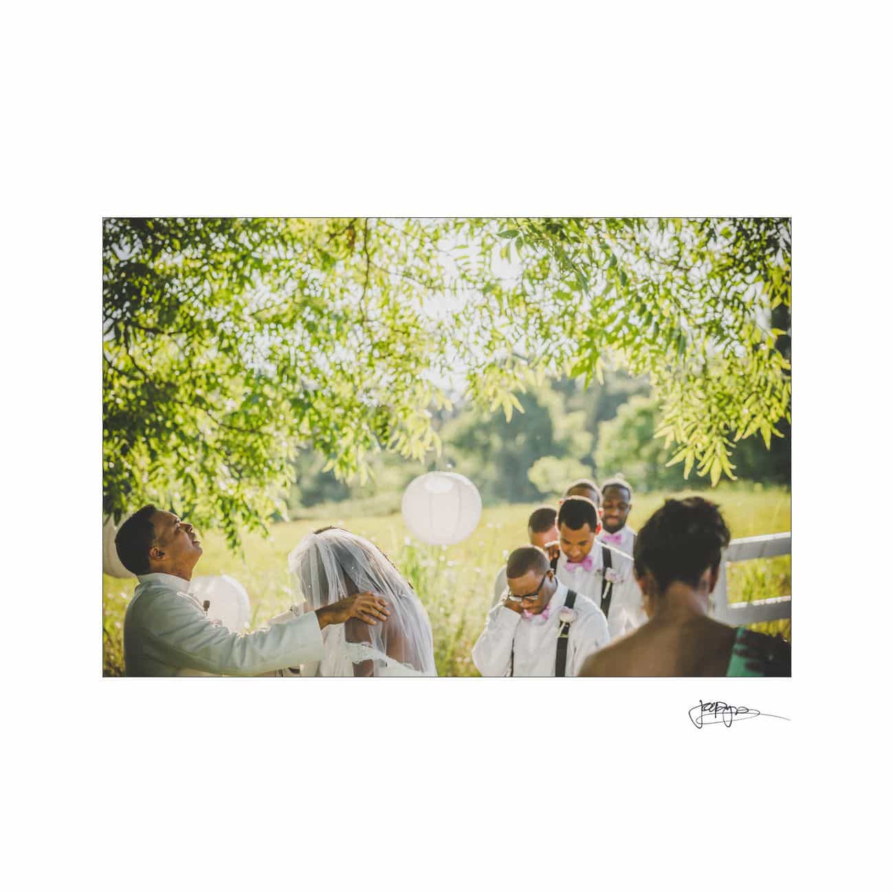 A couple stands under a tree during an outdoor wedding ceremony. The groom, in a white suit, gently holds the brides veil. Guests in formal attire are seated nearby, with lanterns hanging from the tree branches.