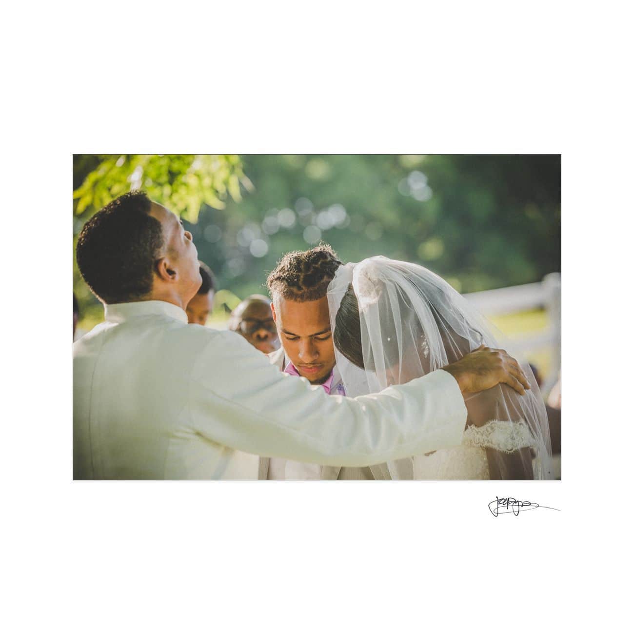 A wedding ceremony scene outdoors with a groom and bride in formal attire, heads bowed together. An officiant in a white suit stands beside them with a hand on the grooms shoulder, under soft natural light.