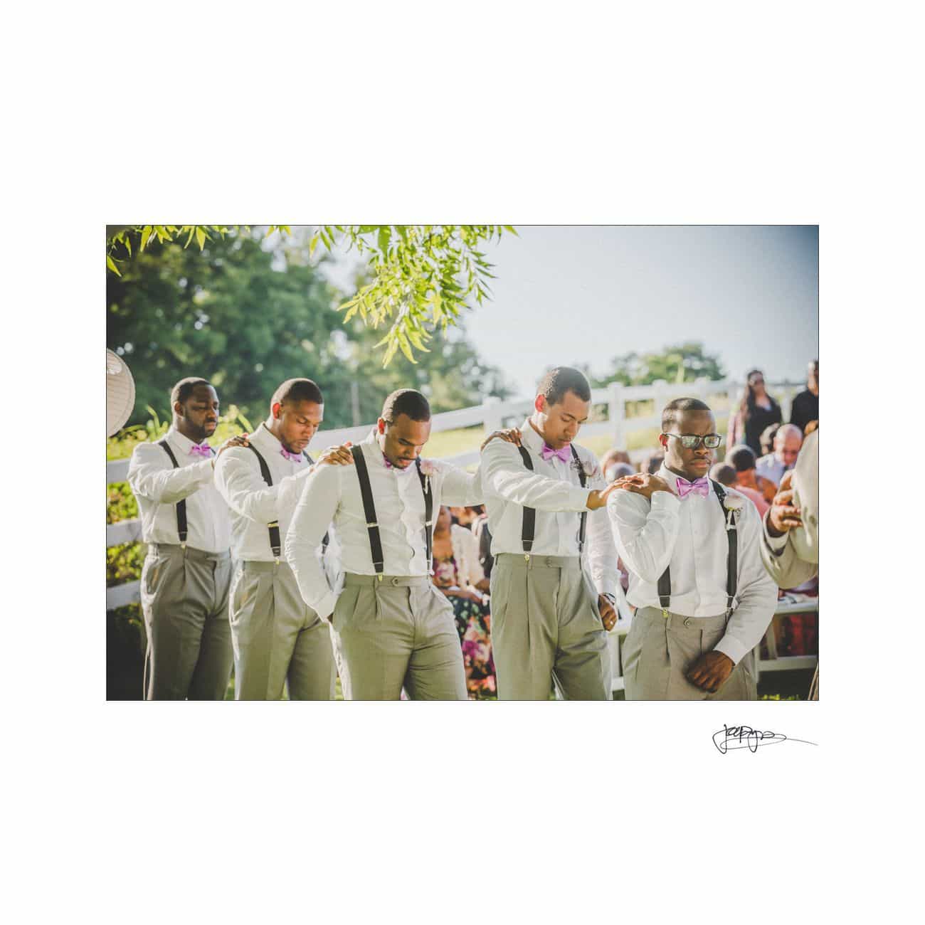 Five groomsmen in white shirts, gray pants, suspenders, and bow ties adjust each others attire outdoors, under a tree. They stand in a line at a wedding ceremony, with guests seated in the background.