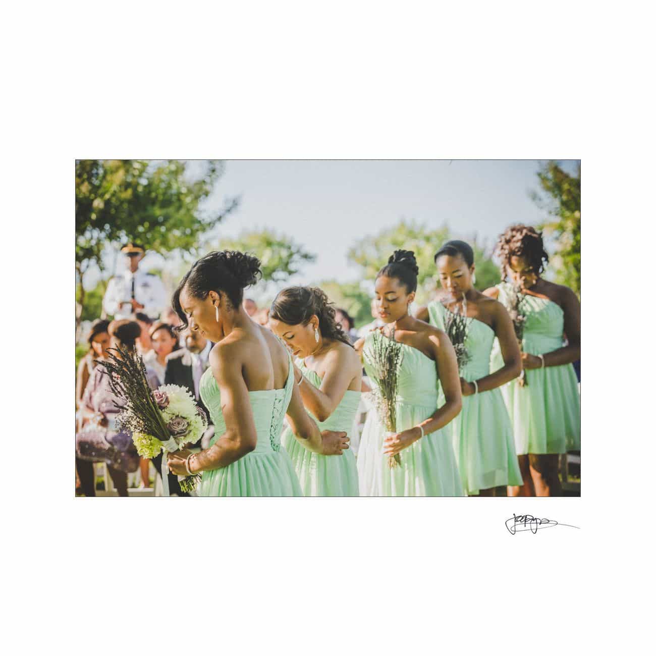 A group of bridesmaids in matching light green dresses hold bouquets and stand in line outdoors during a sunny wedding ceremony. Guests, including children, are seated in the background.