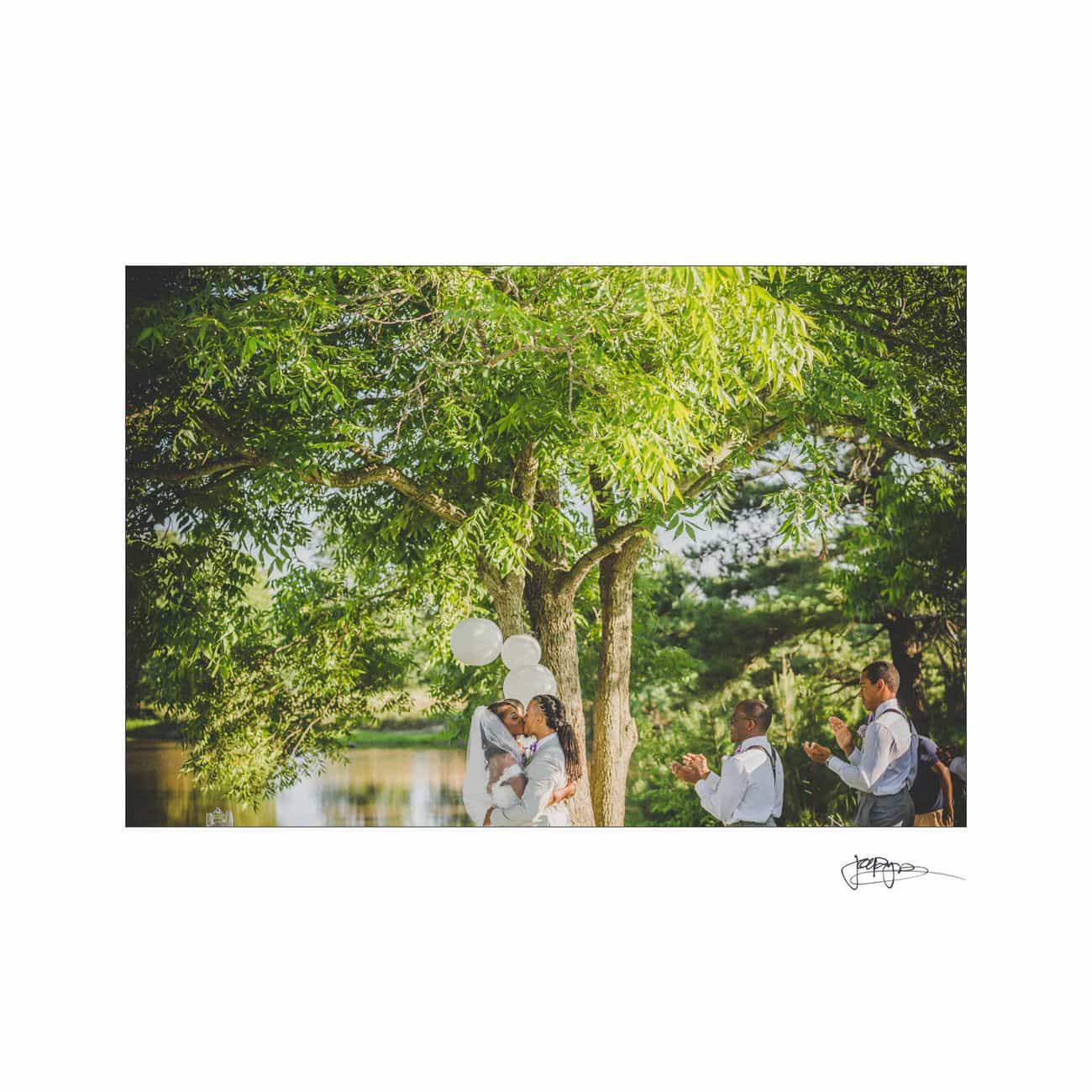 A couple kisses under a tree during a wedding ceremony by a lake. Two people stand nearby, applauding. White balloons are tied to the tree, and the scene is bathed in soft sunlight, creating a serene and romantic atmosphere.