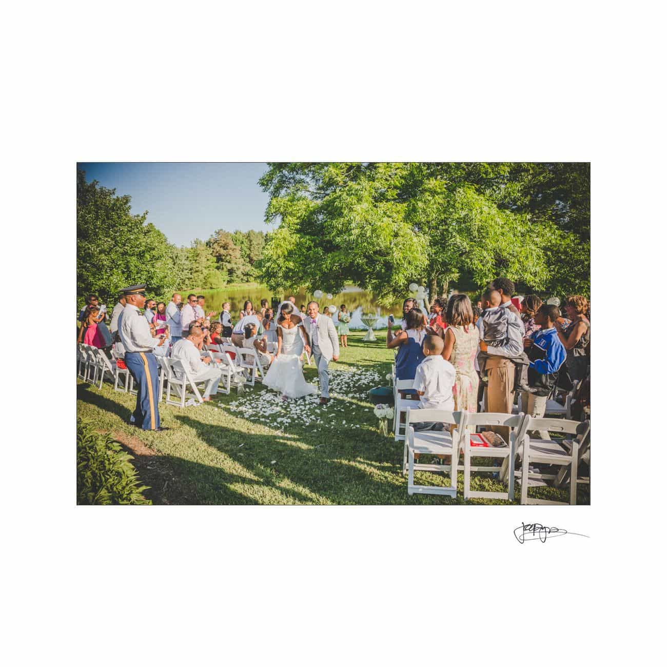A newlywed couple joyfully walks down the outdoor aisle, surrounded by guests on white chairs. The grassy area is adorned with flower petals, and trees provide a lush, green backdrop under a sunny sky.