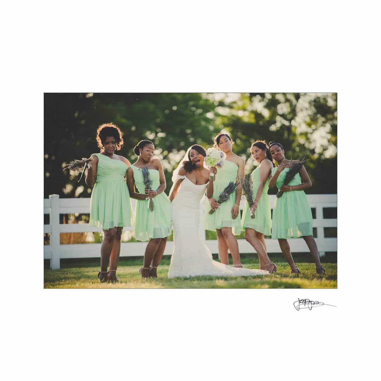 A bride in a white gown and five bridesmaids in light green dresses pose playfully outdoors by a white fence. The bridesmaids hold bouquets, and the scene is set against a backdrop of green trees.