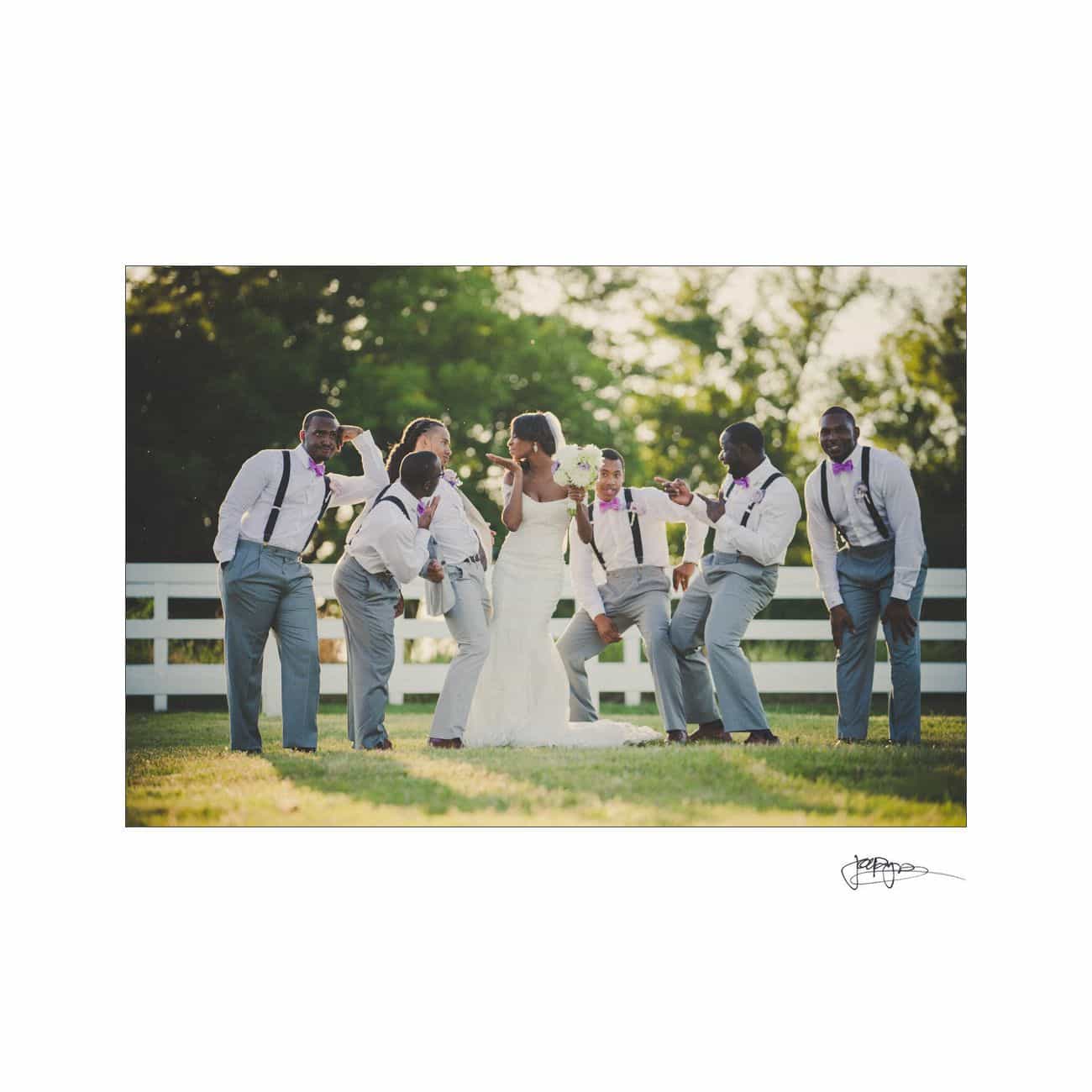 A bride and groom kiss outdoors, surrounded by five groomsmen in gray pants, white shirts, and suspenders. The groomsmen strike playful poses against a backdrop of trees and a white fence.