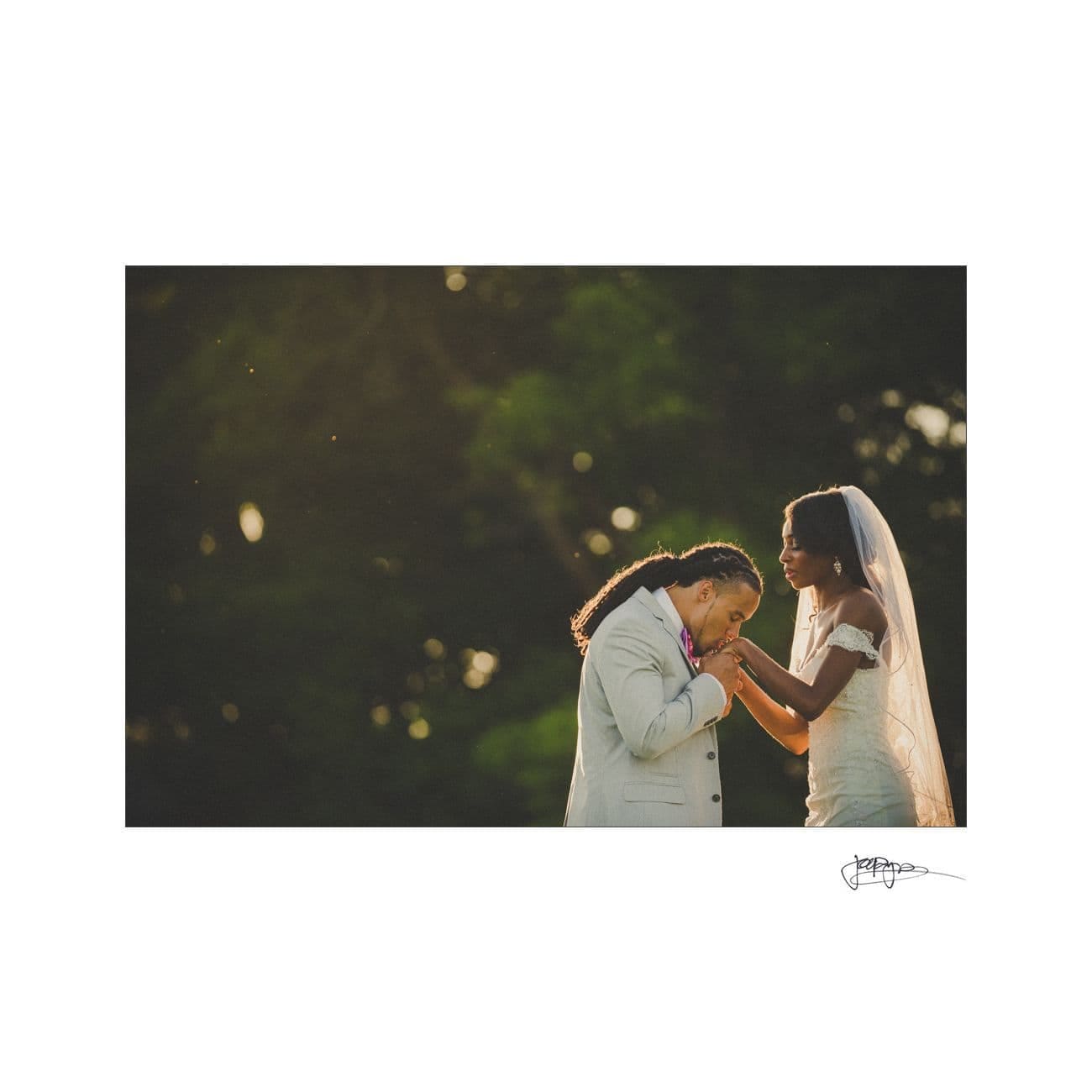 A bride and groom stand outdoors in front of a blurred forest background. The groom, wearing a light gray suit, gently kisses the brides hand. The bride, in a white dress and veil, smiles softly at him.