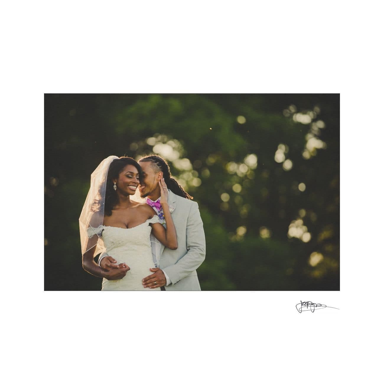 A bride in a white gown and veil smiles while a groom in a light-colored suit embraces her from behind. They stand outdoors with greenery in the background, bathed in soft sunlight.