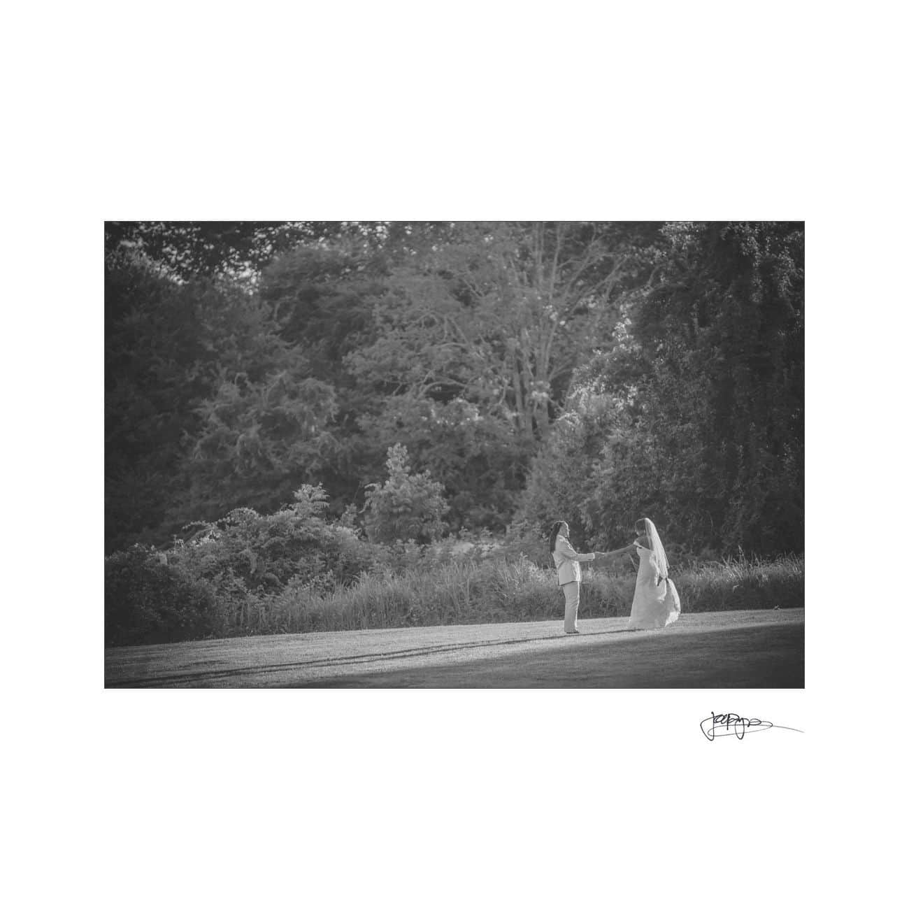 A black and white photo of a bride in a veil and a person holding hands in a grassy field, surrounded by dense trees. A signature is visible at the bottom right corner.