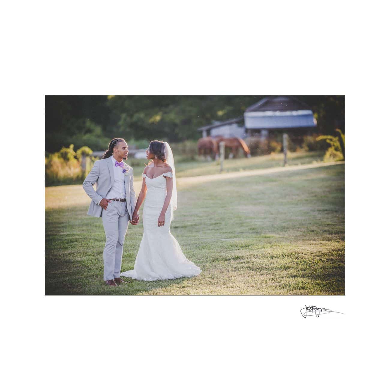 A bride and groom stand together on a grassy field, holding hands. She wears a white gown and veil, while he wears a light gray suit with a red bow tie. A blurred background includes a rustic shed and two horses.