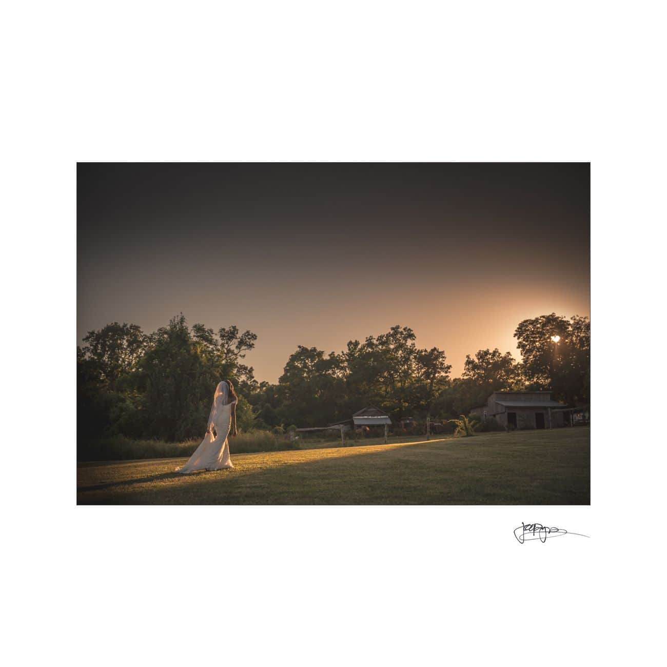 A bride in a long white dress walks on a path through a grassy field at sunset. Trees and a house are visible in the background. The sky has a warm, golden hue.
