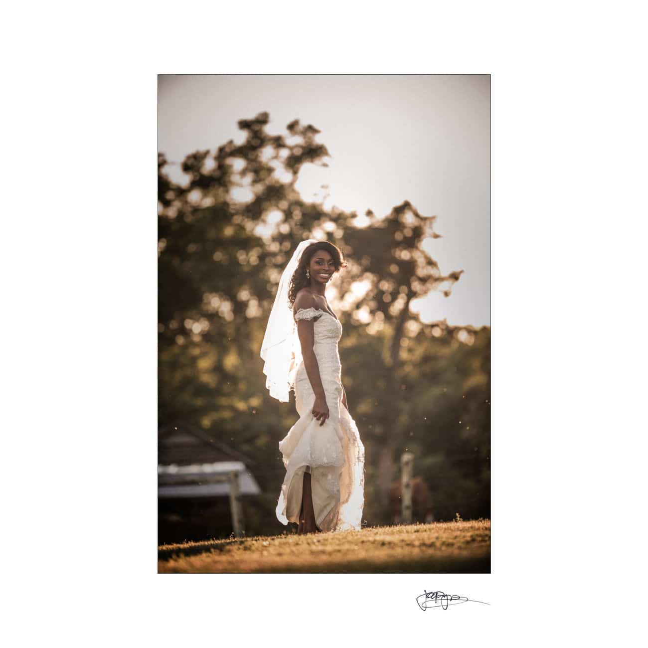 A bride in a white dress with a veil smiles while standing outdoors on a grassy hill. Sunlight filters through the trees in the background, creating a warm and glowing atmosphere.