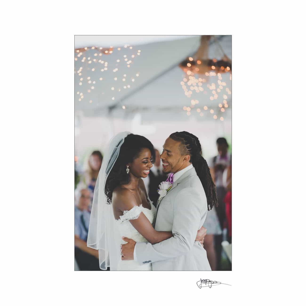 A bride and groom share a joyful dance under a tent adorned with string lights. The bride wears an off-shoulder white gown and veil, while the groom is in a light gray suit with a purple boutonnière. Guests are visible in the blurred background.