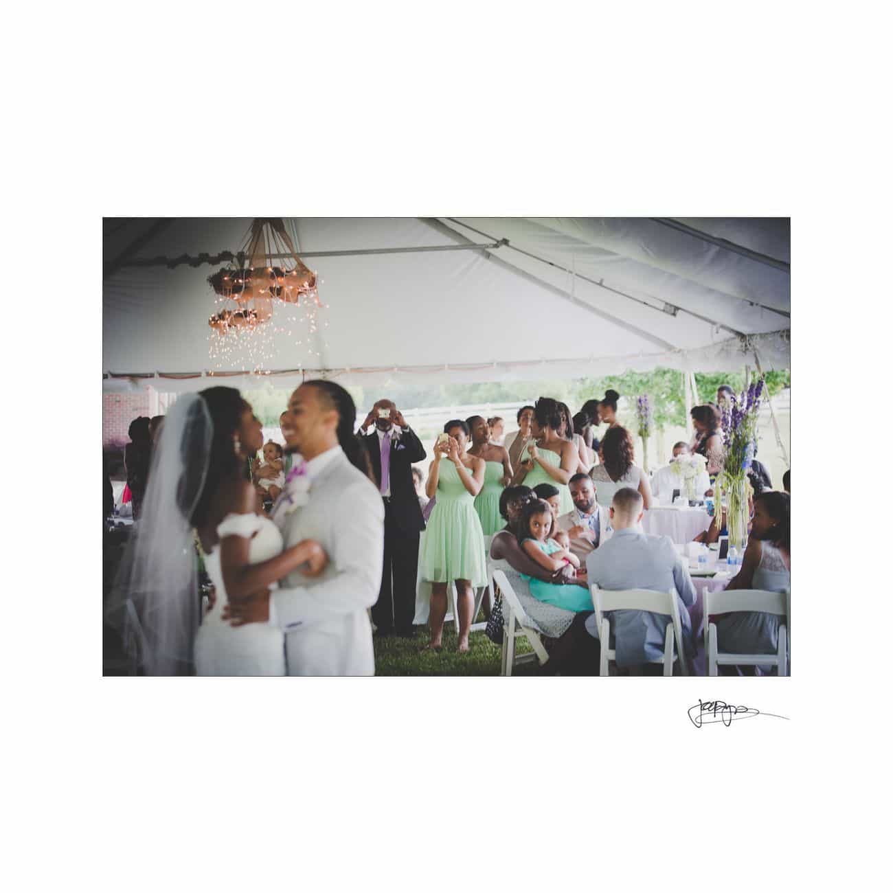 A bride and groom share their first dance under a tent at their outdoor wedding. Guests watch and take photos, while some are seated at tables decorated with white tablecloths. The atmosphere is festive and joyful.