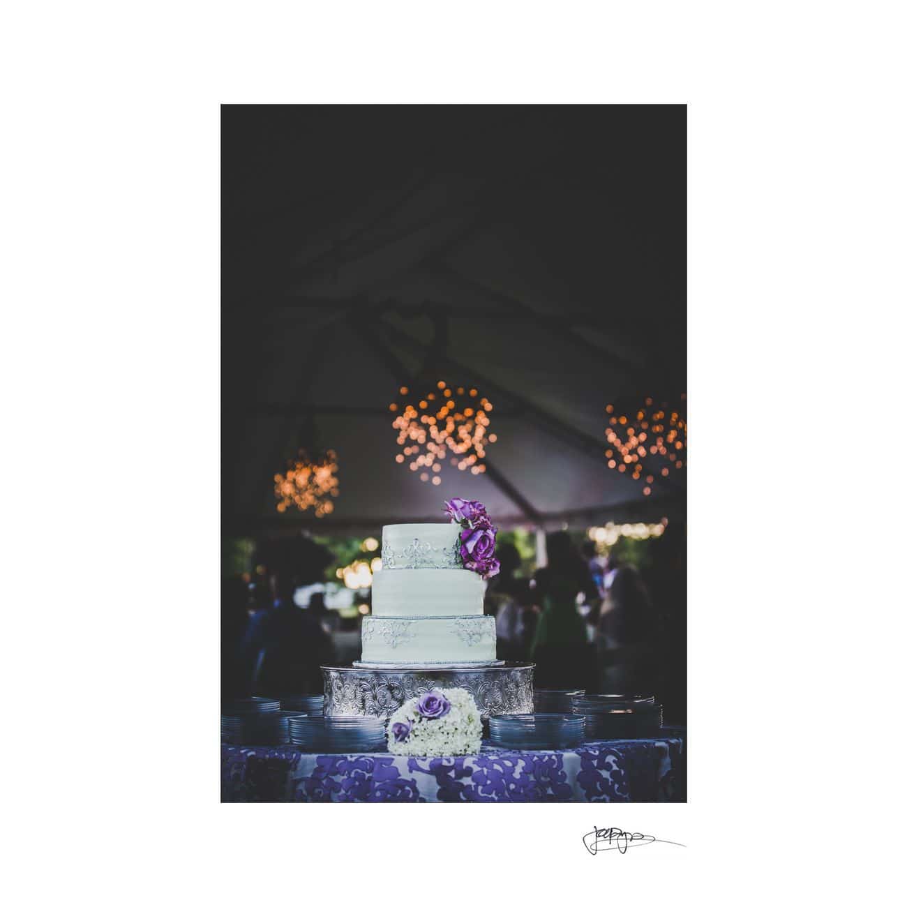 A three-tier white wedding cake with purple flower decorations is displayed under a tent. The background features soft, glowing lights creating a warm ambiance. The cake sits on a silver stand with a bouquet of flowers at its base.