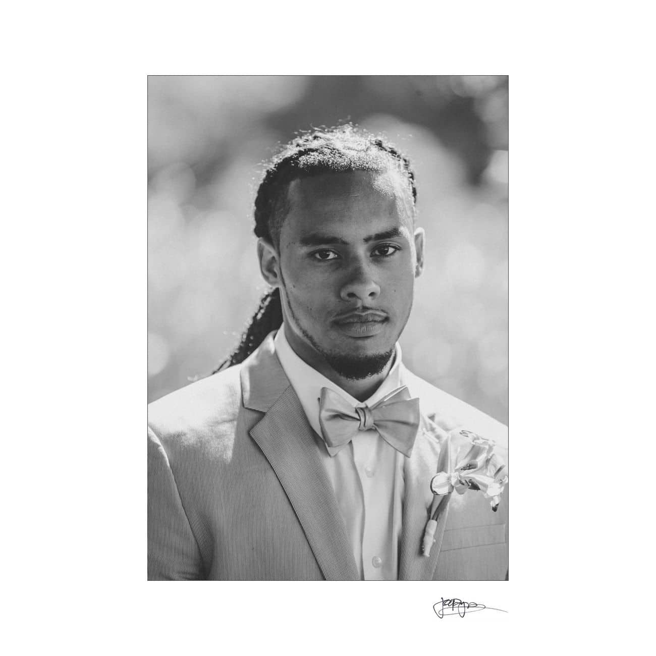 Black and white portrait of a man wearing a suit, bow tie, and floral boutonniere, with braided hair. The background is softly blurred, and the image has an elegant, formal composition.